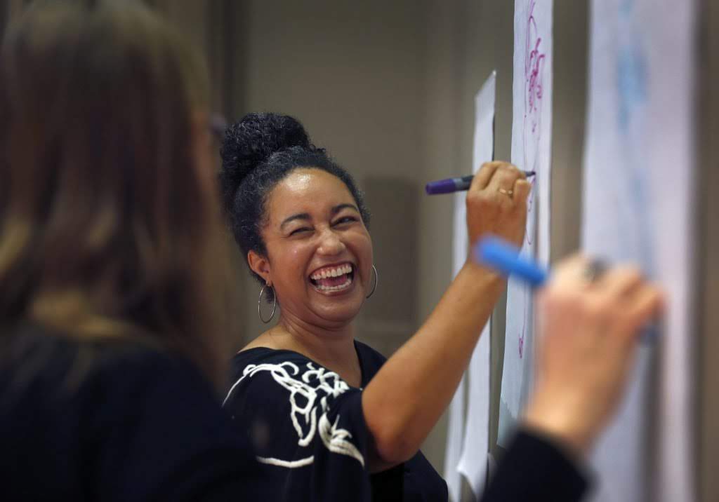 Two women smiling and drawing on paper with markers during a workshop or collaborative event.