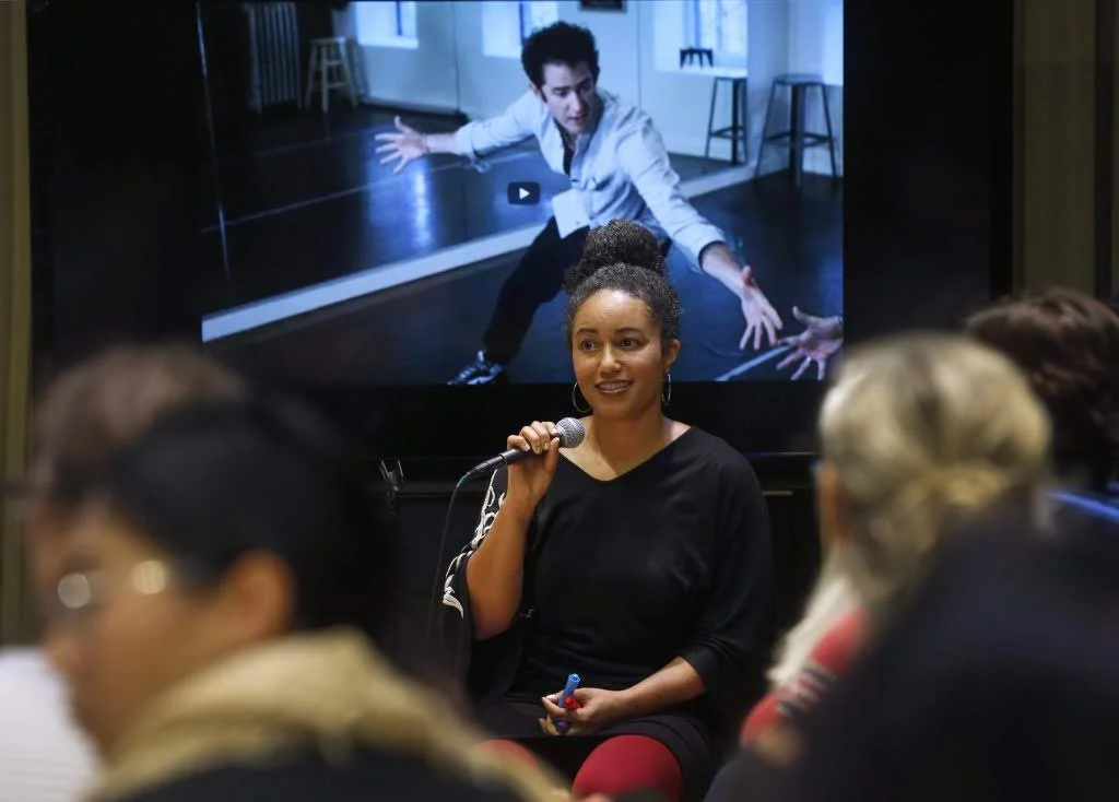 A woman holding a microphone sits in front of a TV screen displaying a dance video. She is engaged in a conversation with an audience. The screen behind her shows a man mid-movement in a dance studio.