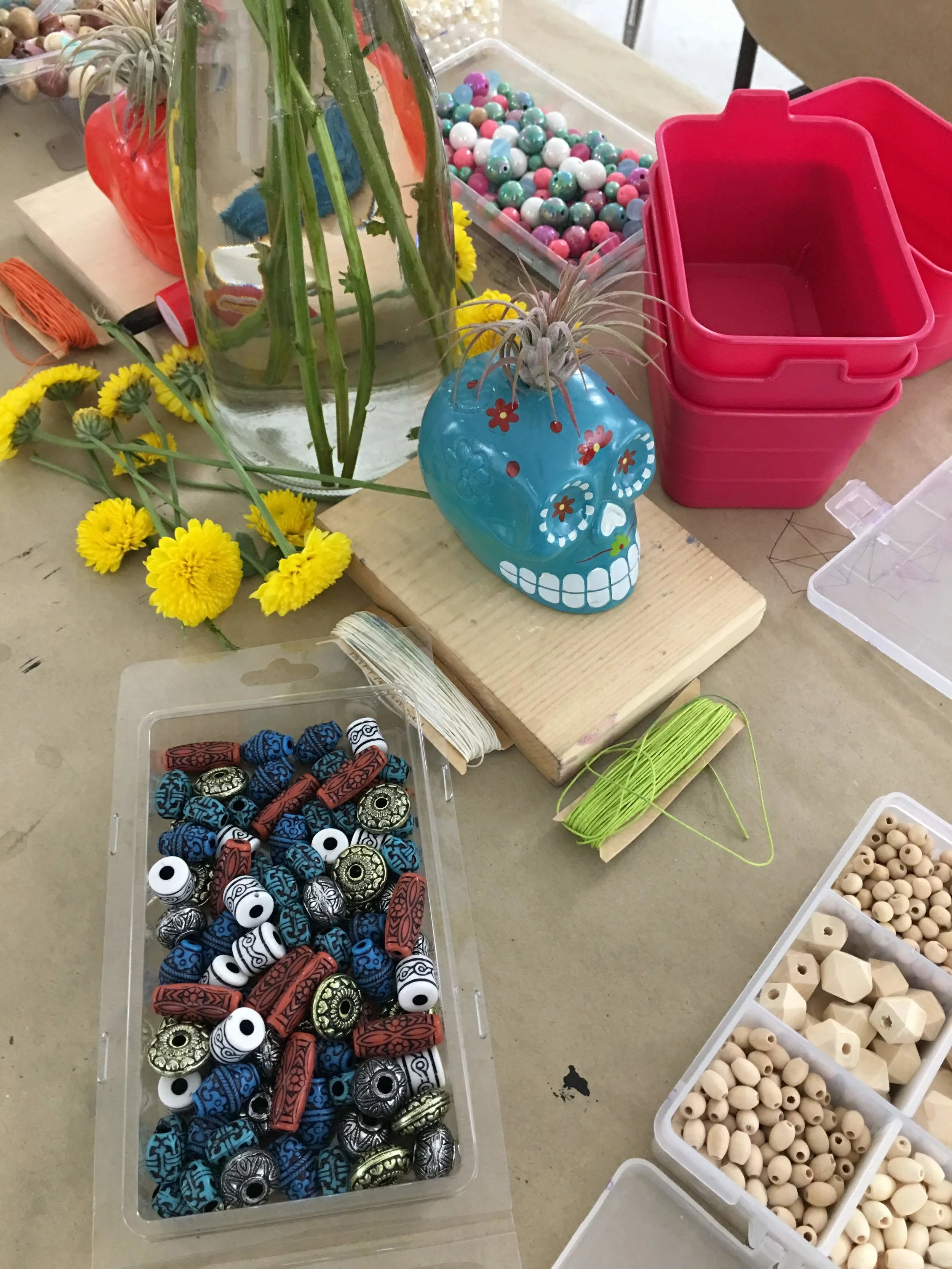 Beads and craft supplies on a table, including colorful skull planters, small yellow flowers, and containers of various beads and string.