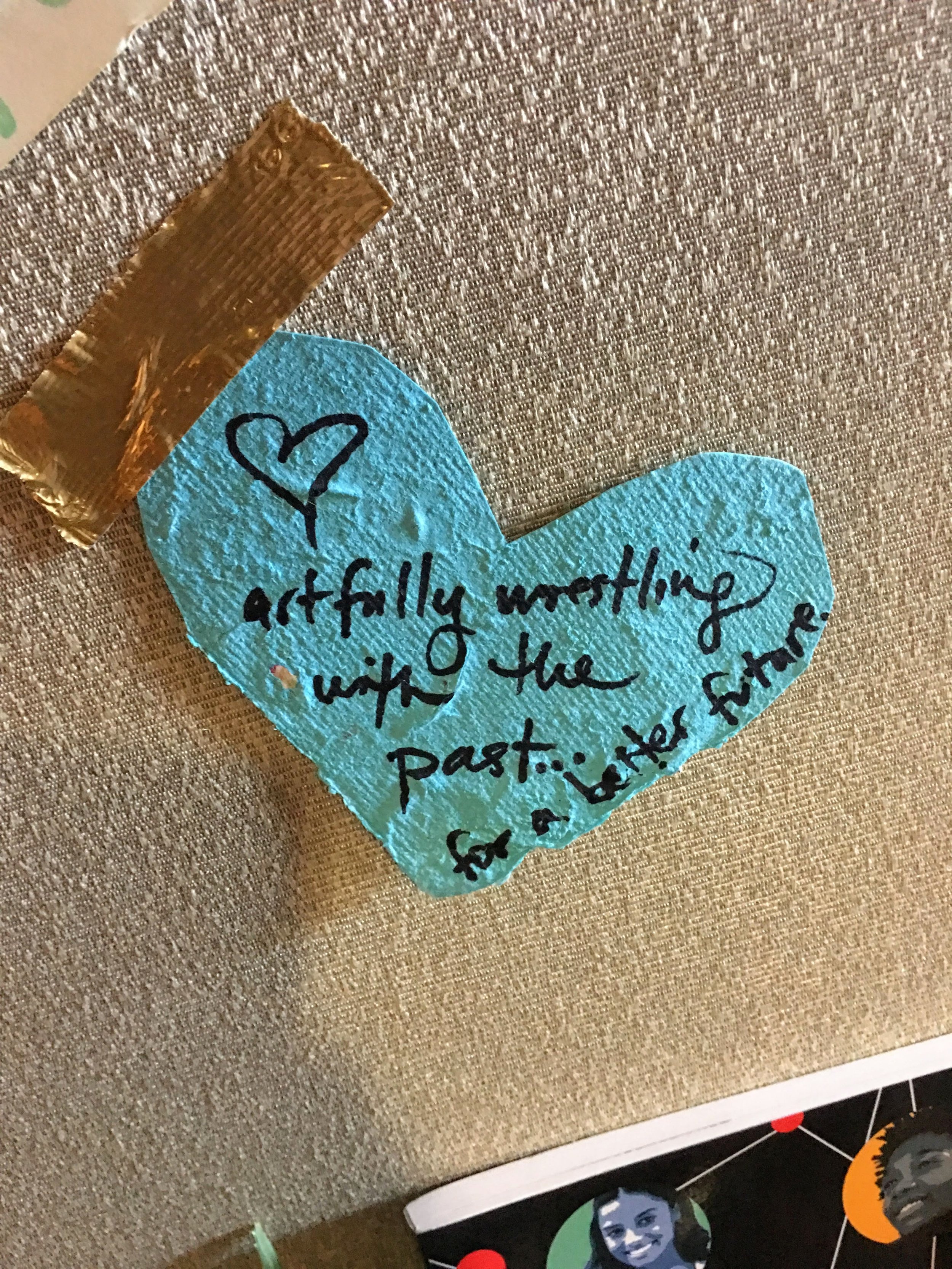 Handwritten note on blue heart shape with quote: "artfully wrestling with the past... for a better future," attached to a fabric board with gold tape.