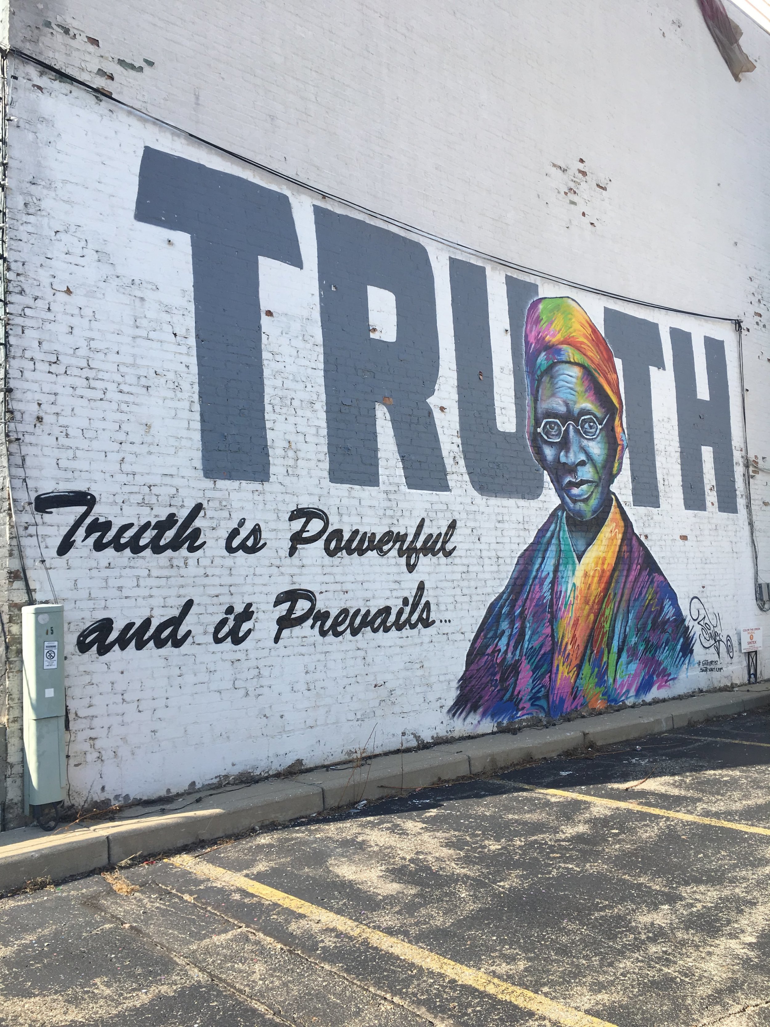 Mural on a white brick wall featuring large gray letters spelling "TRUTH," with a colorful portrait of a woman wearing a headscarf. Below the word, it reads "Truth is Powerful and it Prevails."