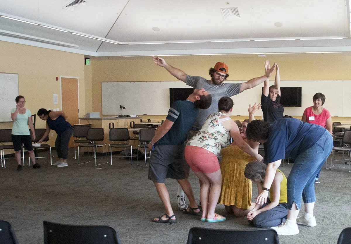 Group of people participating in a team-building exercise in a room with chairs and whiteboards.