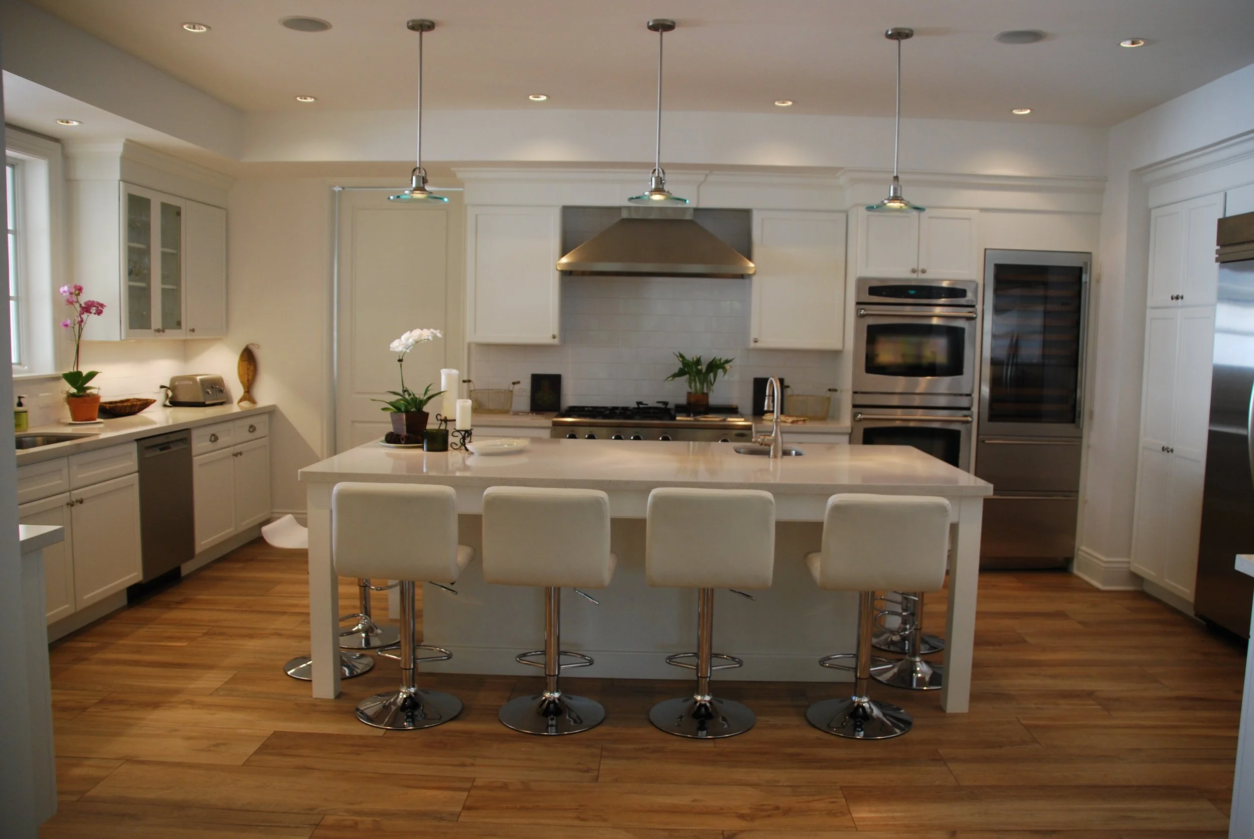 Highland Beach white kitchen with a large island, upholstered counter stools, stainless hood, and warm wood flooring