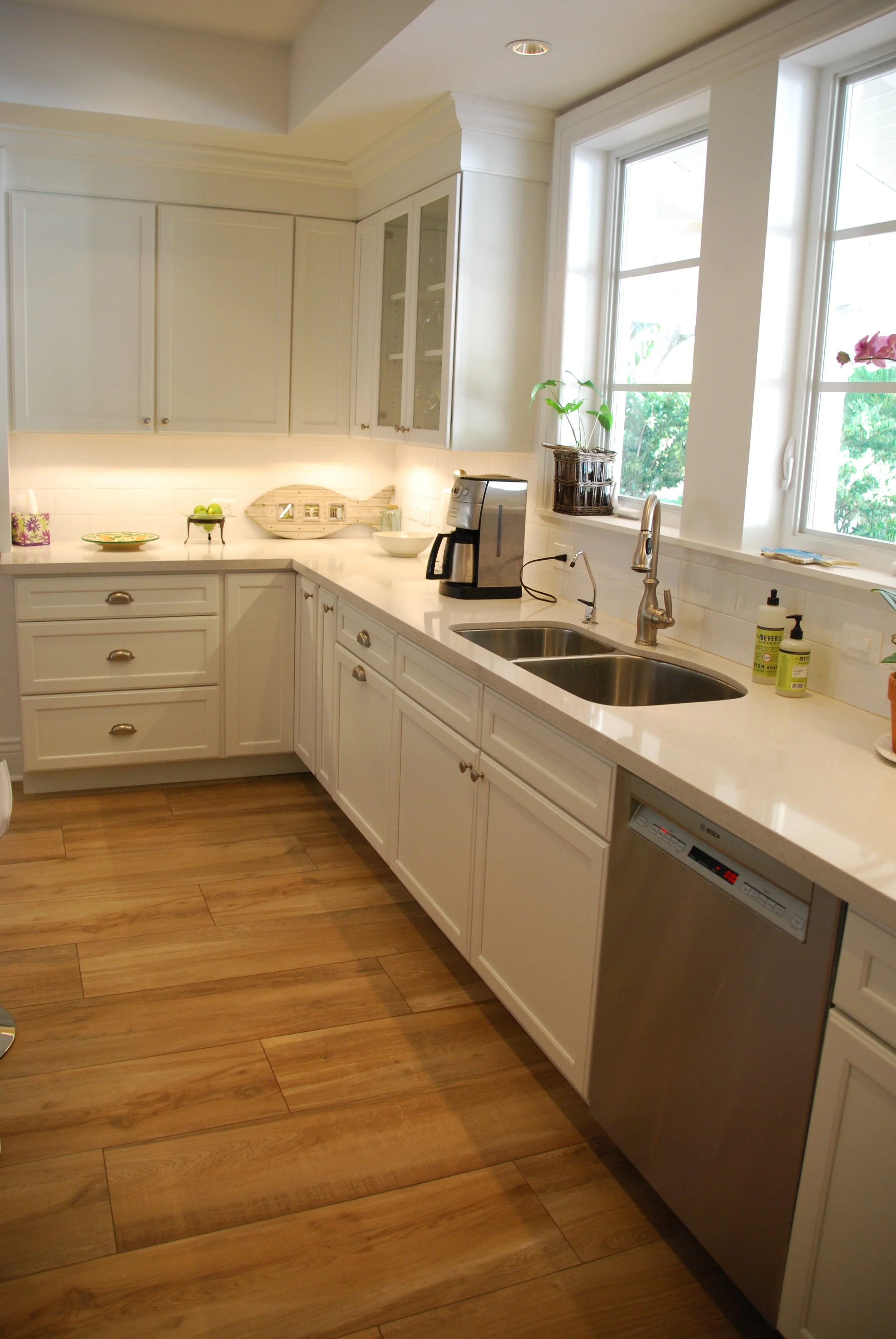 White sink cabinetry beneath a kitchen window with a dishwasher, glass-front upper cabinets, and undercabinet lighting