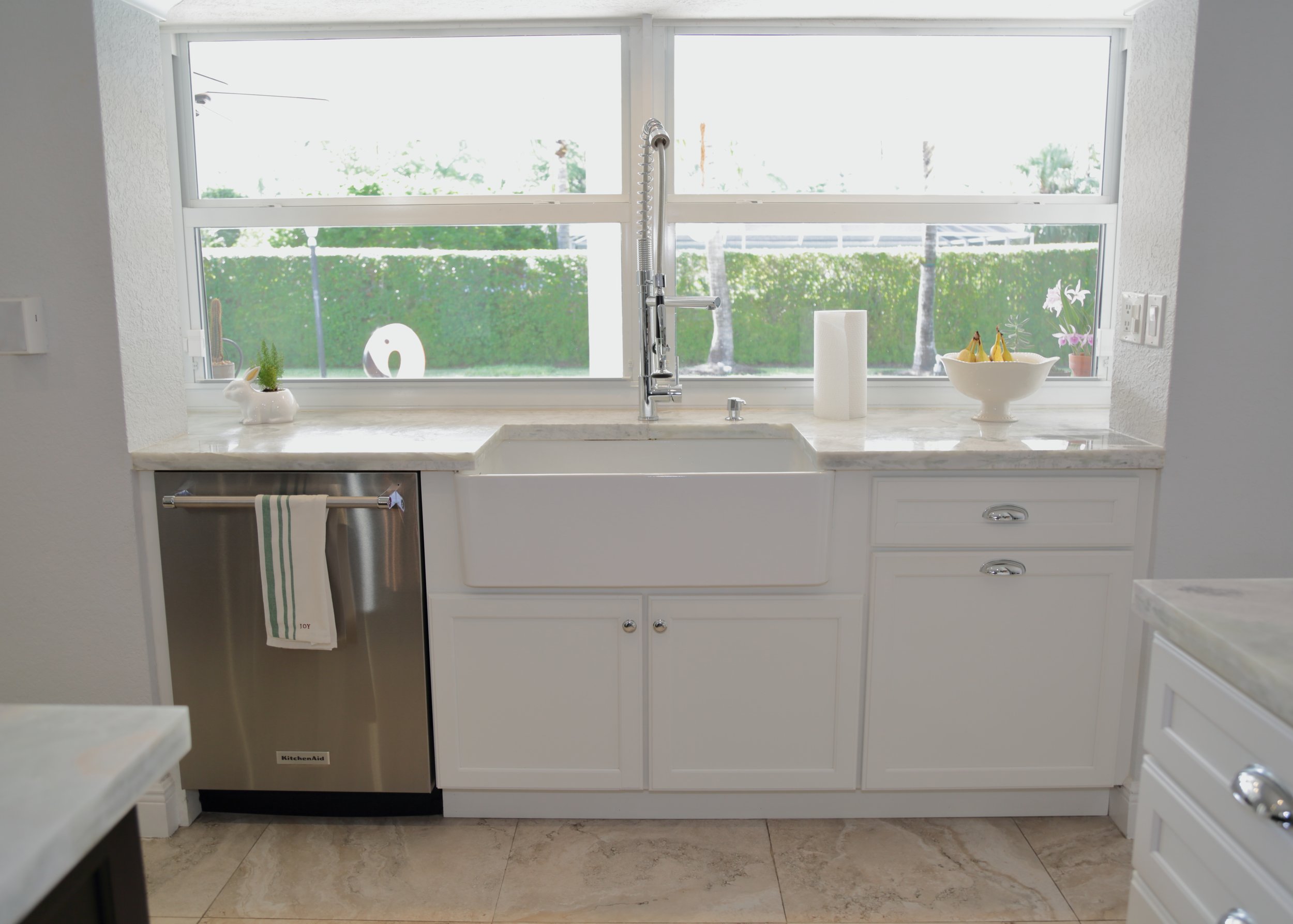 White apron-front sink beneath kitchen windows with drawer cabinets and a stainless dishwasher