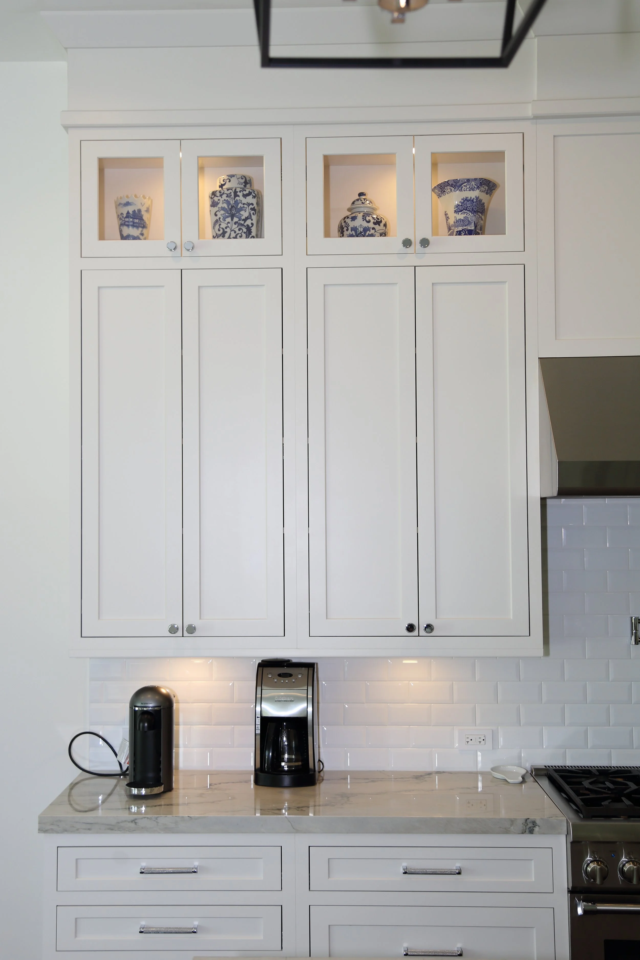 White kitchen coffee station with drawer storage, upper cabinets, and illuminated glass-front display doors