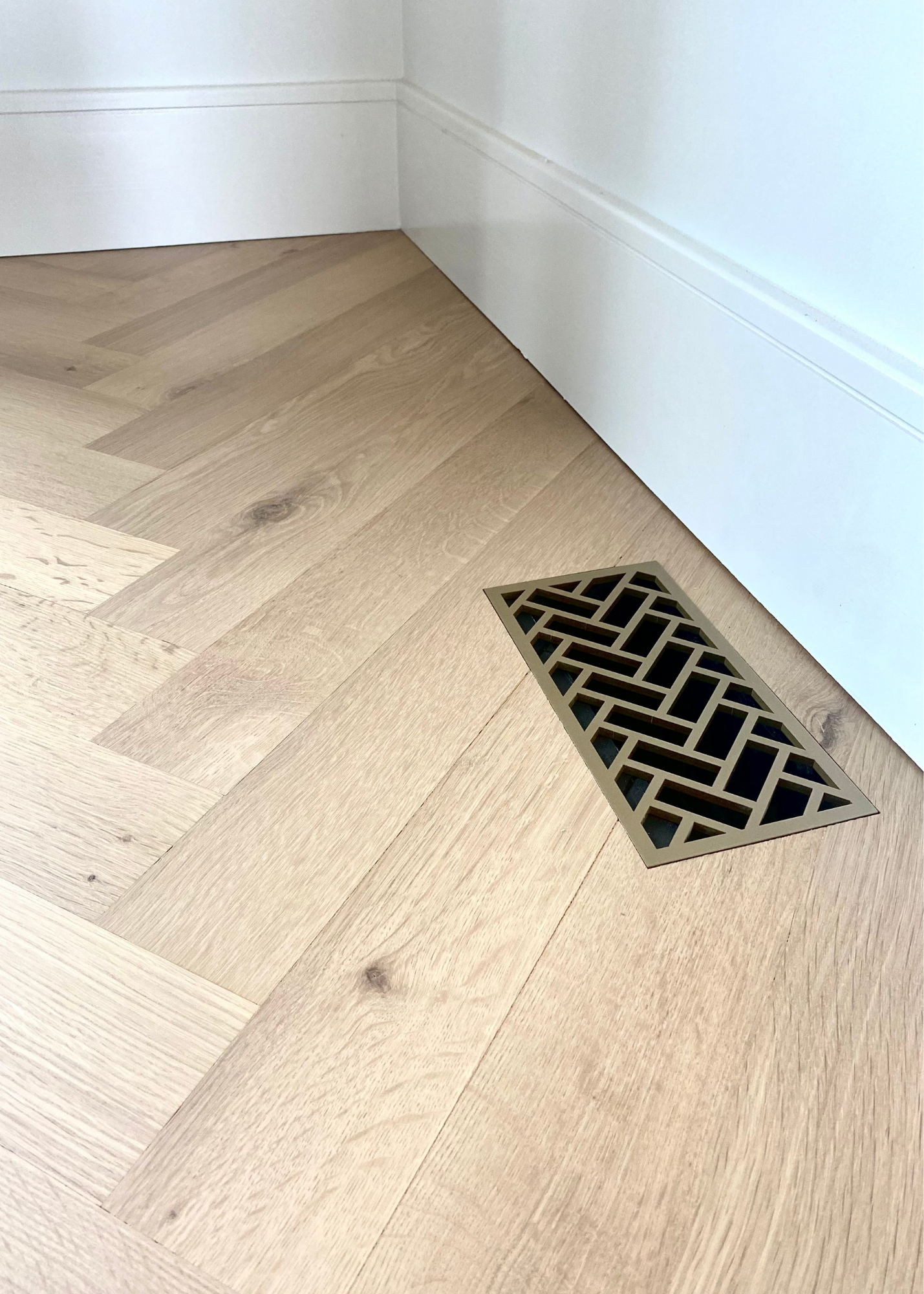 Decorative floor grill flush mounted into a white oak herringbone floor. This steel flush mount vent has a herringbone design and is powder coated vintage gold. 