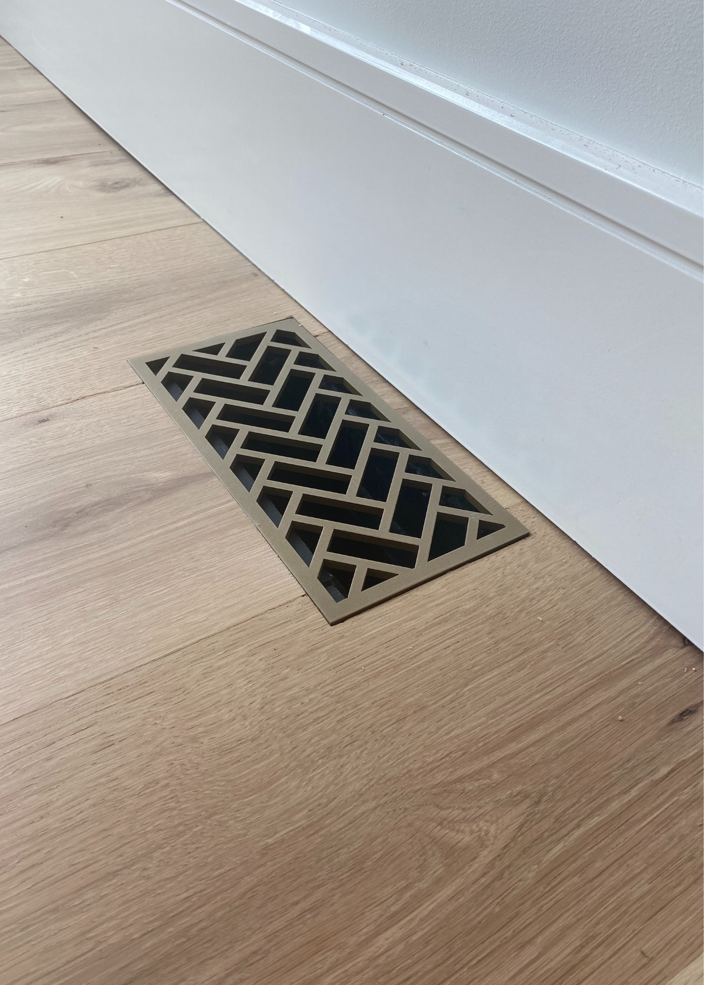 Flush mount floor vent installed into white oak flooring. This decorative grill has a herringbone design and is powder coated vintage gold. 
