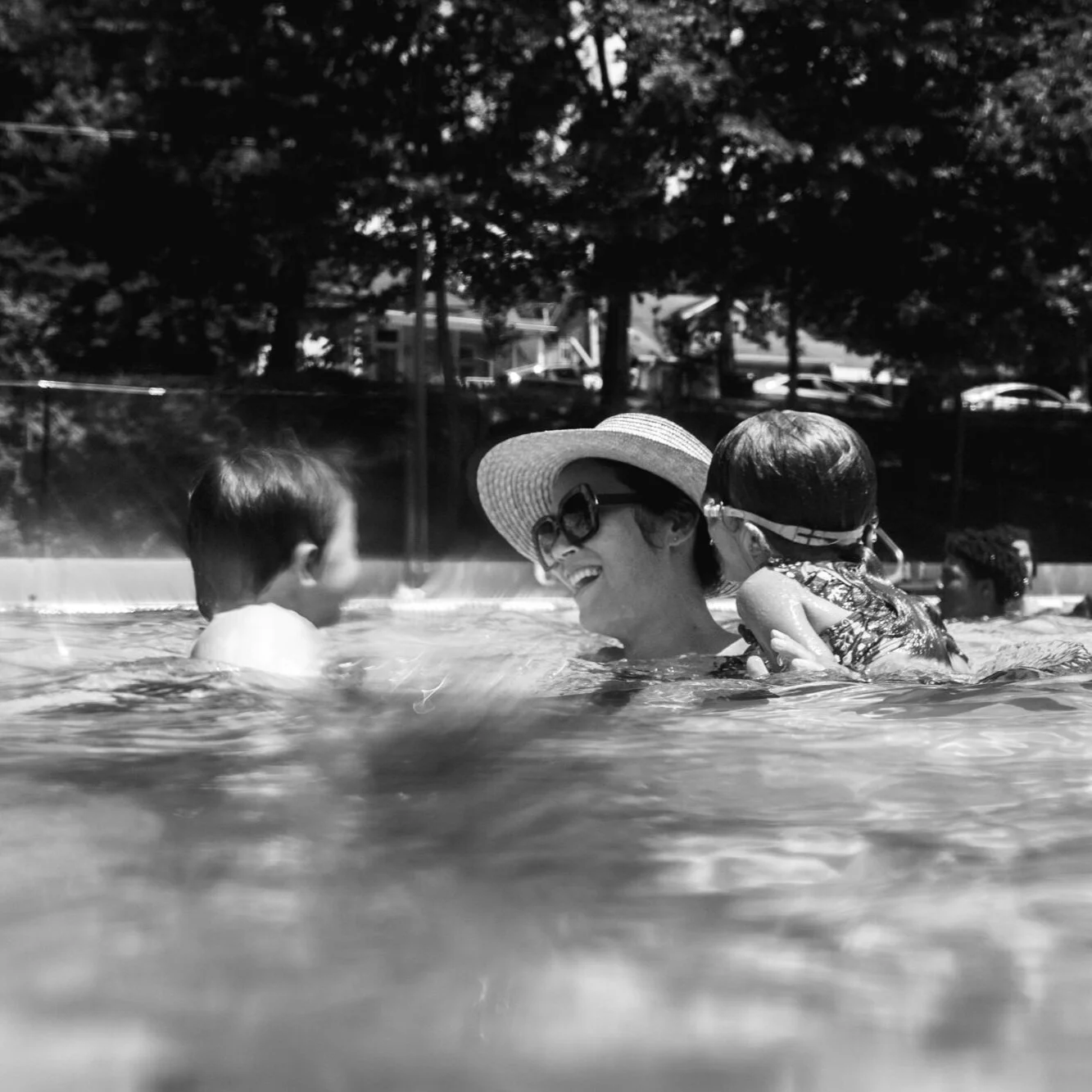image of a mother smiles at her son in a public pool while her daughter hangs on her shoulder from a day in the life photography session