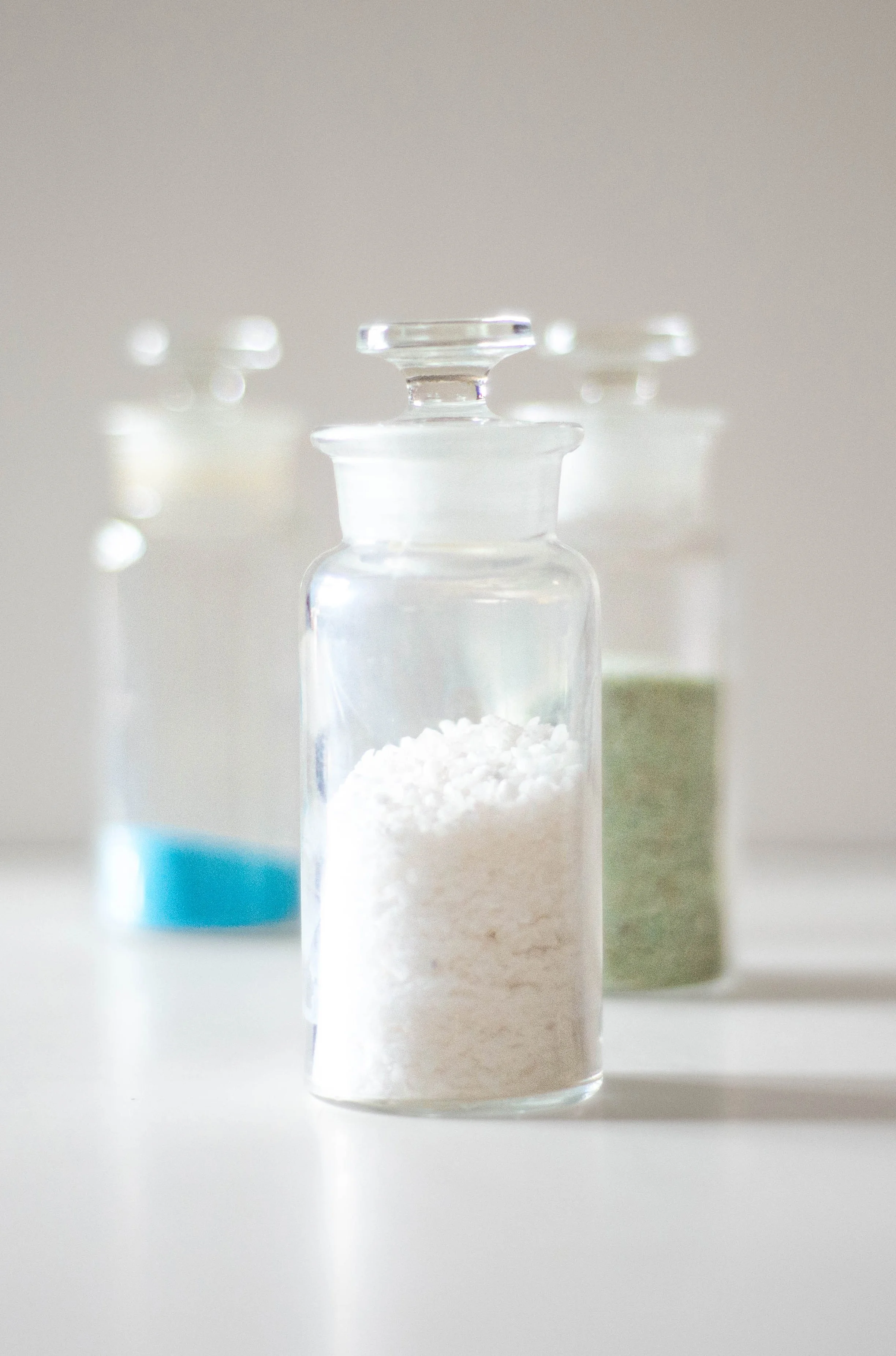 three stoppered glass jars containing crystalline powders sit on a white surface