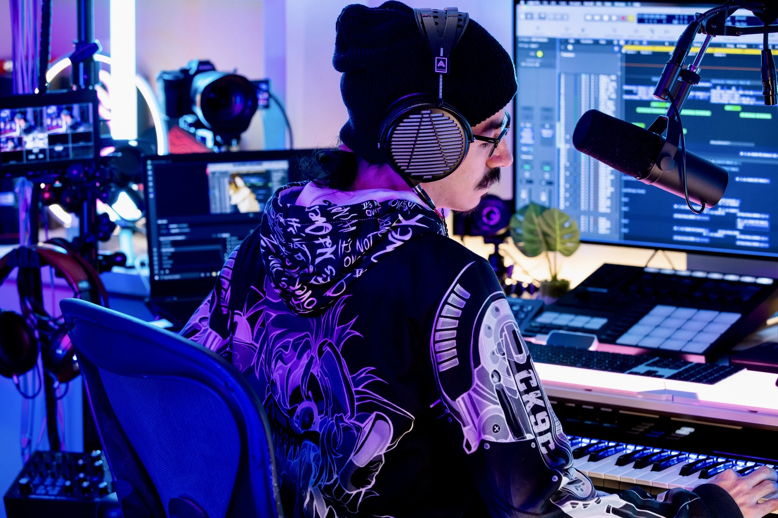 A person in headphones and glasses working on music production in a studio with multiple computer screens and music equipment, illuminated by colorful neon lights.