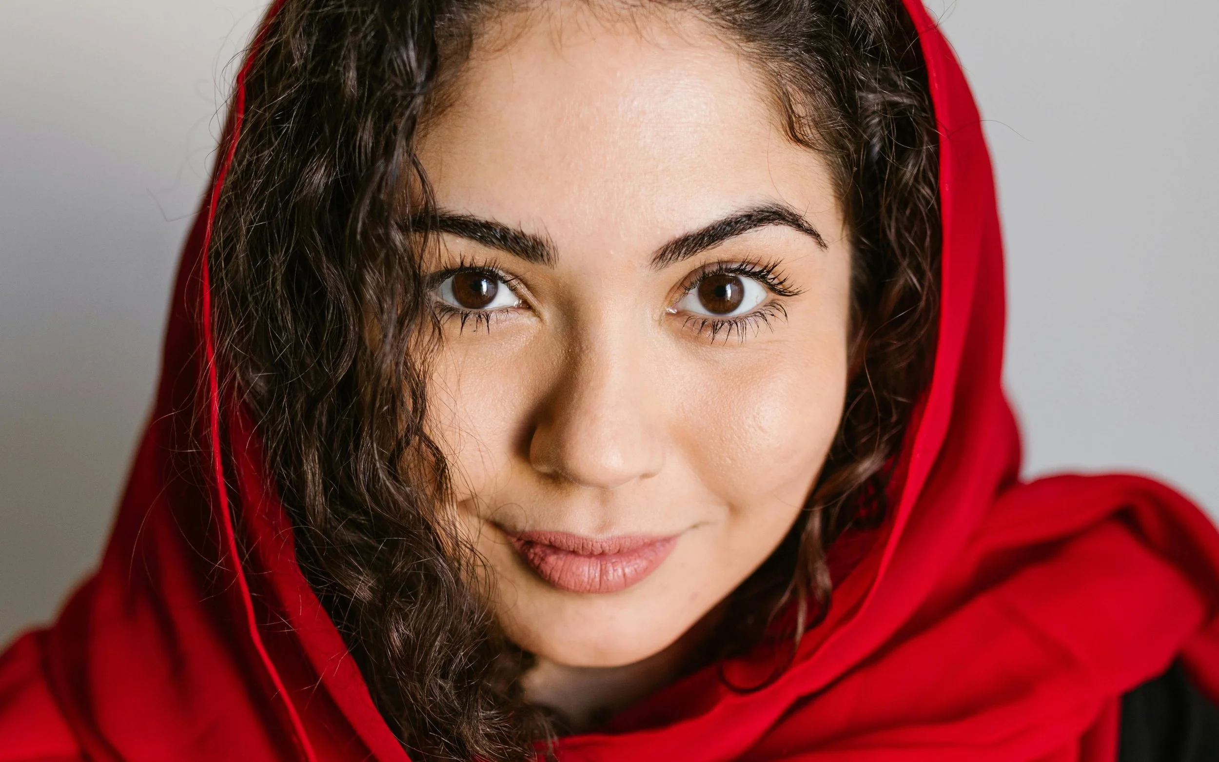 Close-up of a woman with dark curly hair wearing a red hoodie with excellent eyelashed, looking directly at the camera with brown eyes and a slight smile.