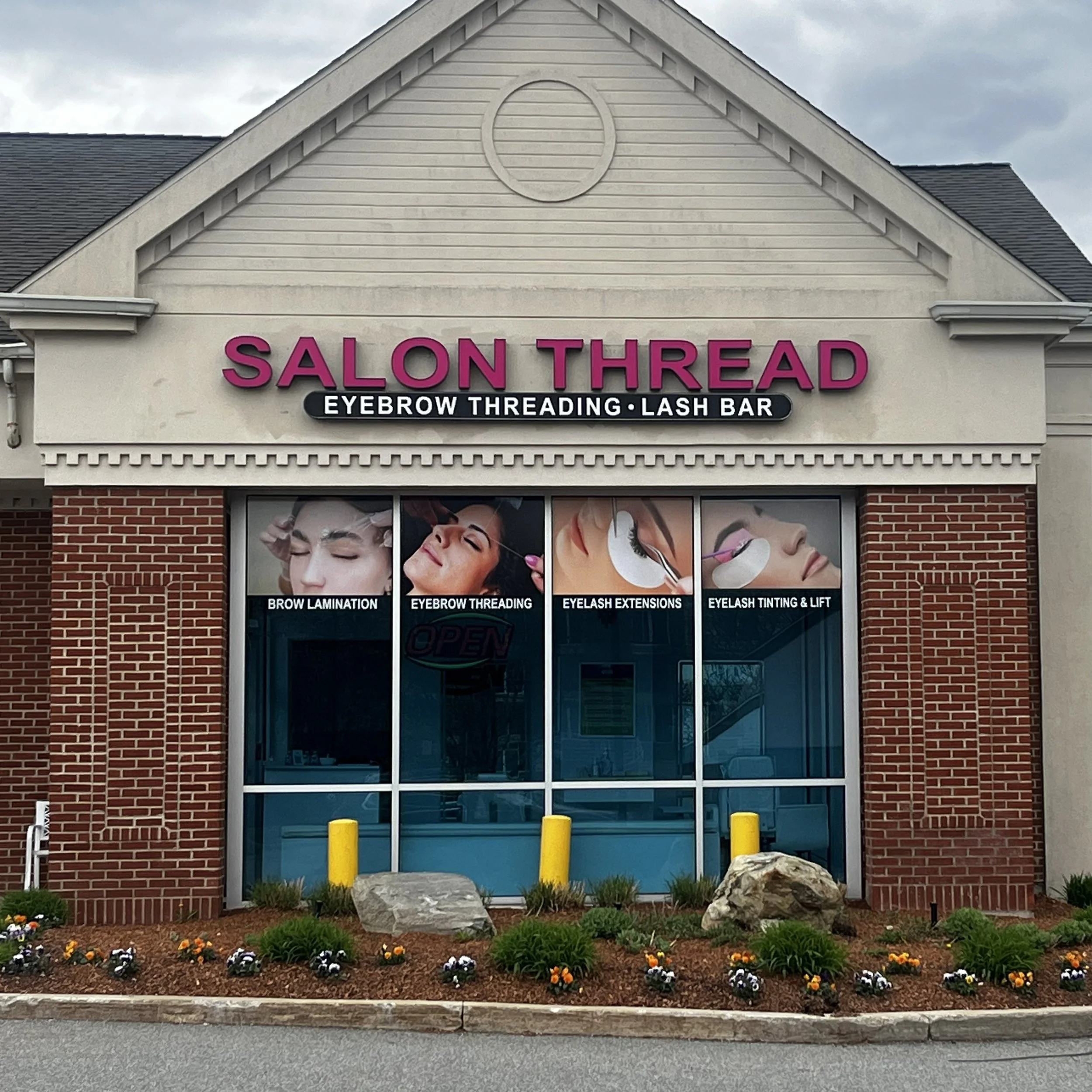front view of a beauty salon with a sign reading 'Salon Thread' and services listed including eyebrow threading, lash bar, brow lamination, eyelash extensions, and eyelash tinting and lift. The salon has a brick facade, large windows showing images of women getting beauty treatments, and a small flower bed with colorful flowers and rocks outside.