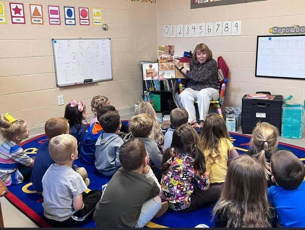 In a classroom, a woman sits in front of a group of students reading them a book.