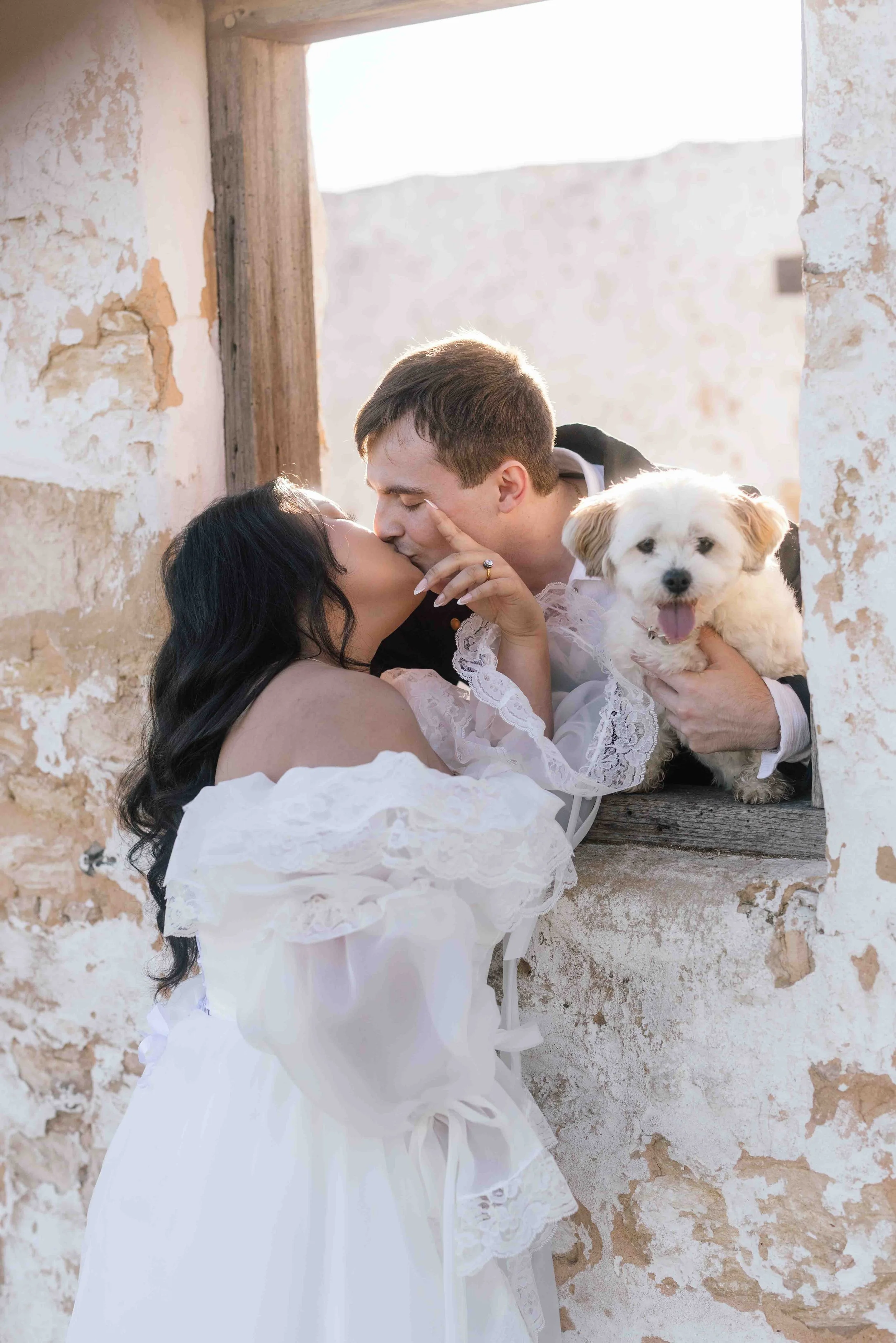 Bride in off-shoulder lace gown kissing her groom through a weathered stone window at Perry's Paddock, their happy white dog