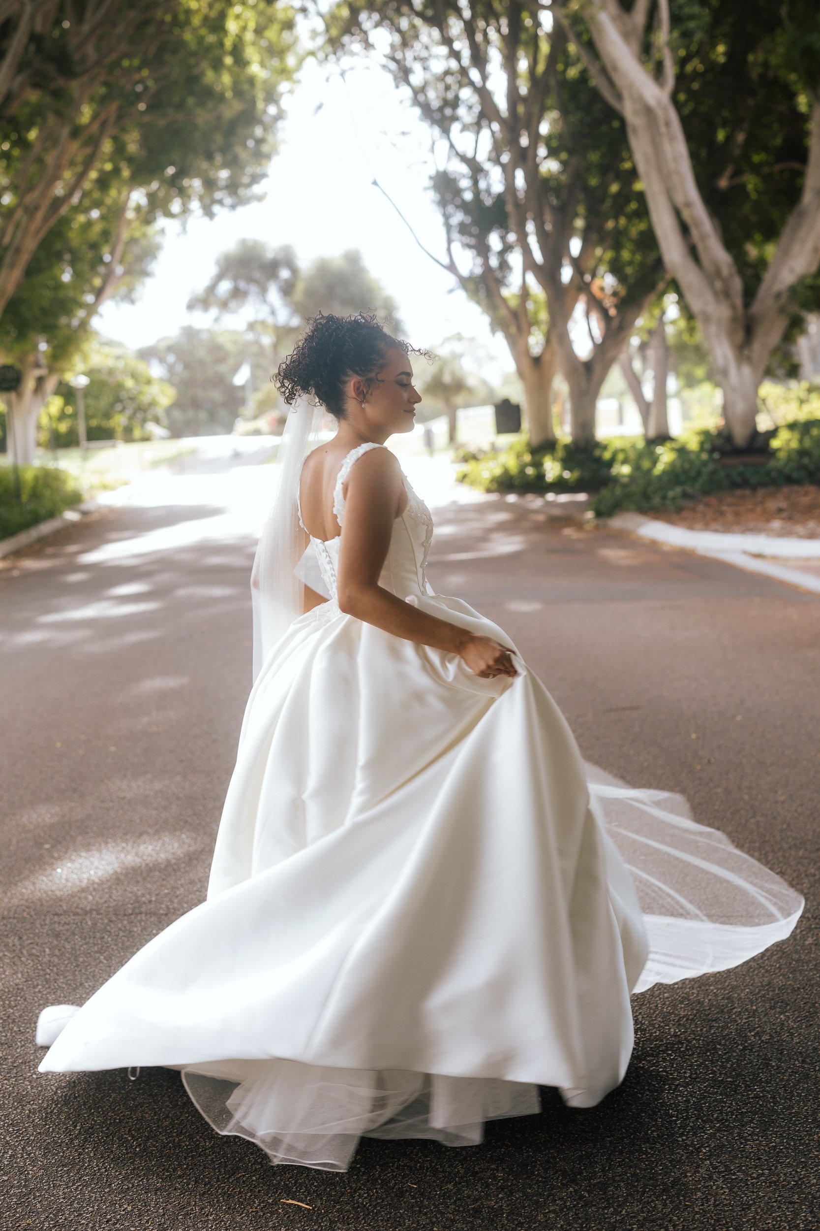 A bride twirls her flowing satin ball gown and veil along a sun-dappled tree-lined driveway, captured by a Perth wedding