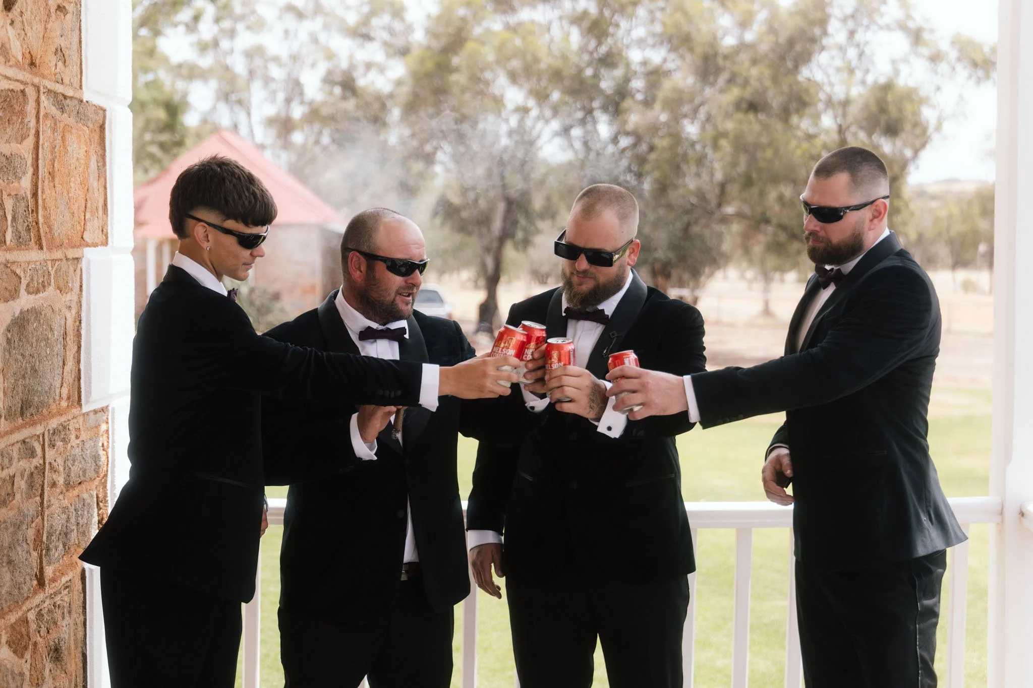 Groomsmen in black velvet tuxedos and sunglasses cheering with drinks on the historic white verandah of Buckland Estate, a premier country wedding venue in the Avon Valley.