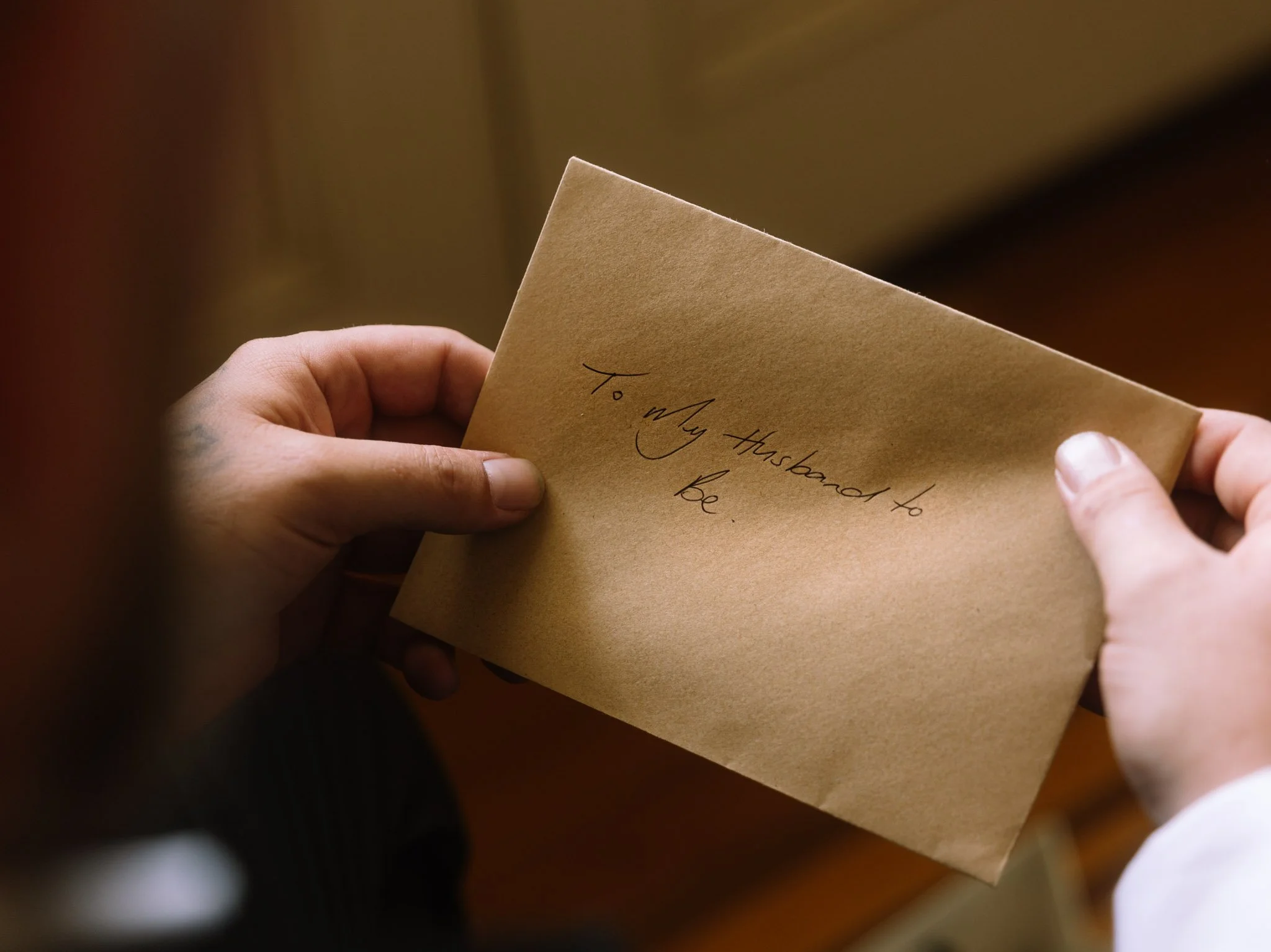 Close-up macro detail of a groom's hands holding a kraft paper envelope with handwritten text reading 'To My husband to be' during morning wedding preparations at Buckland Estate.