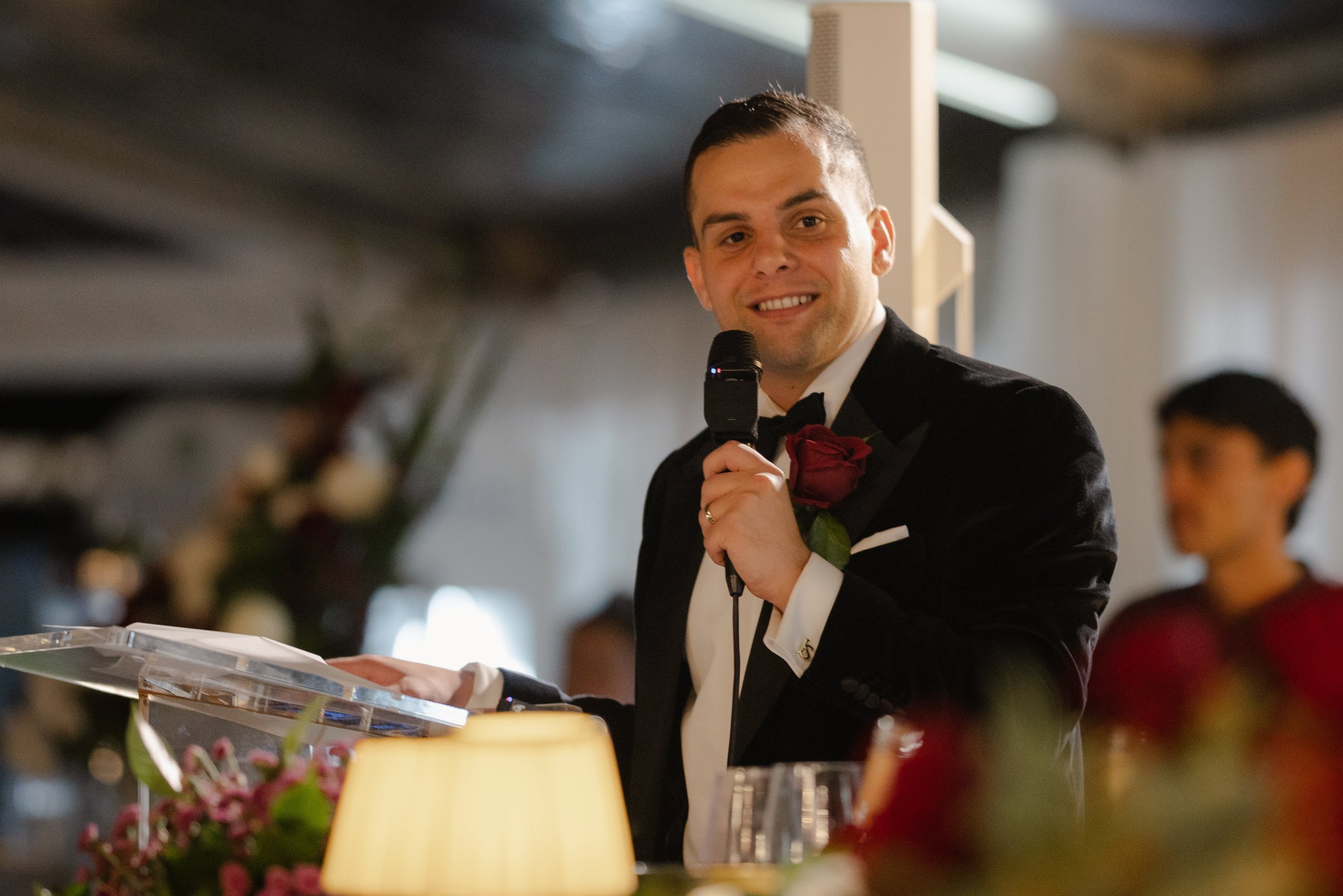 Groom in a black tuxedo and red rose boutonniere smiles warmly while delivering his wedding speech at an acrylic lectern.