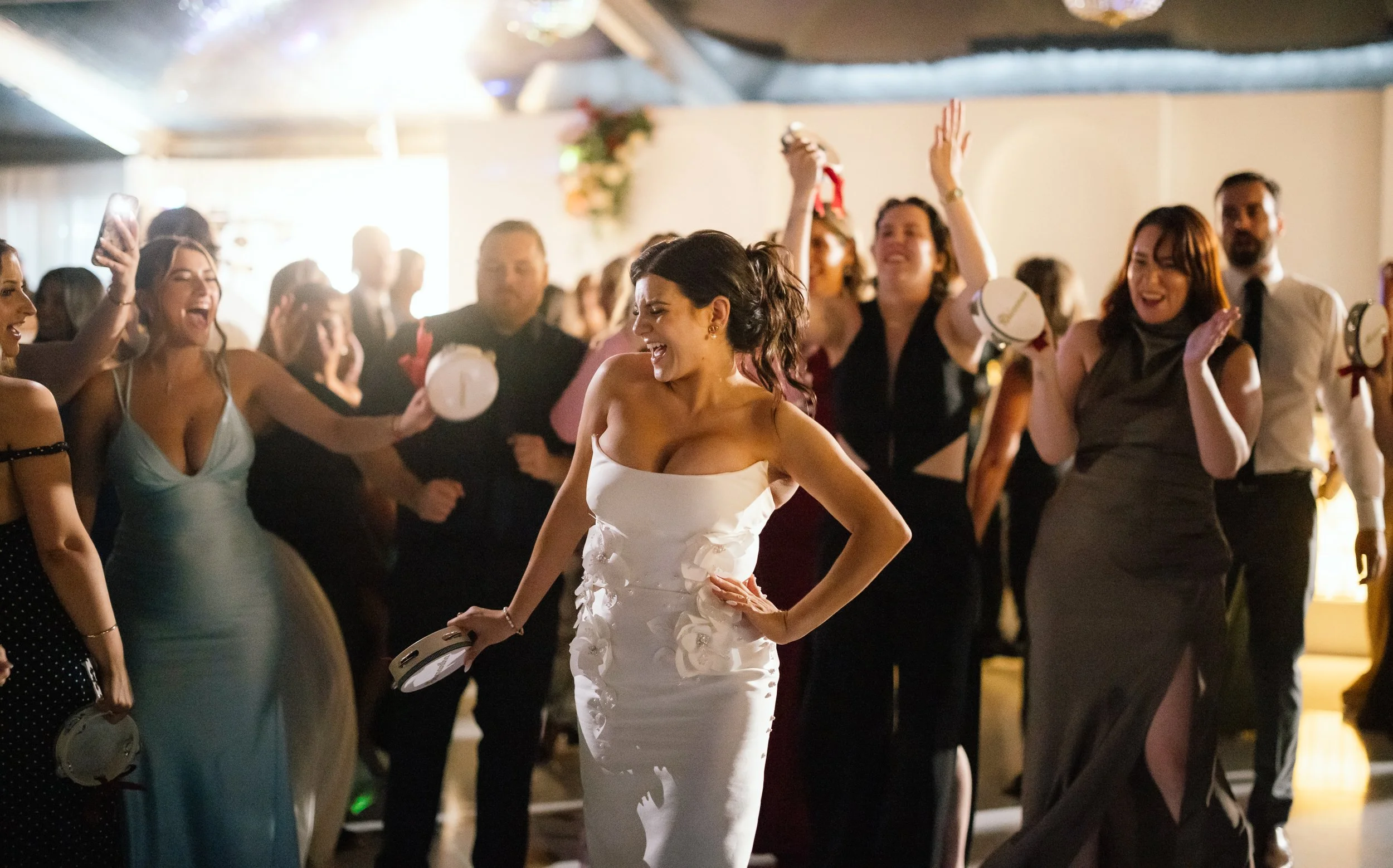 Laughing bride in a fitted floral appliqué gown dances with a tambourine as jubilant guests celebrate on the reception