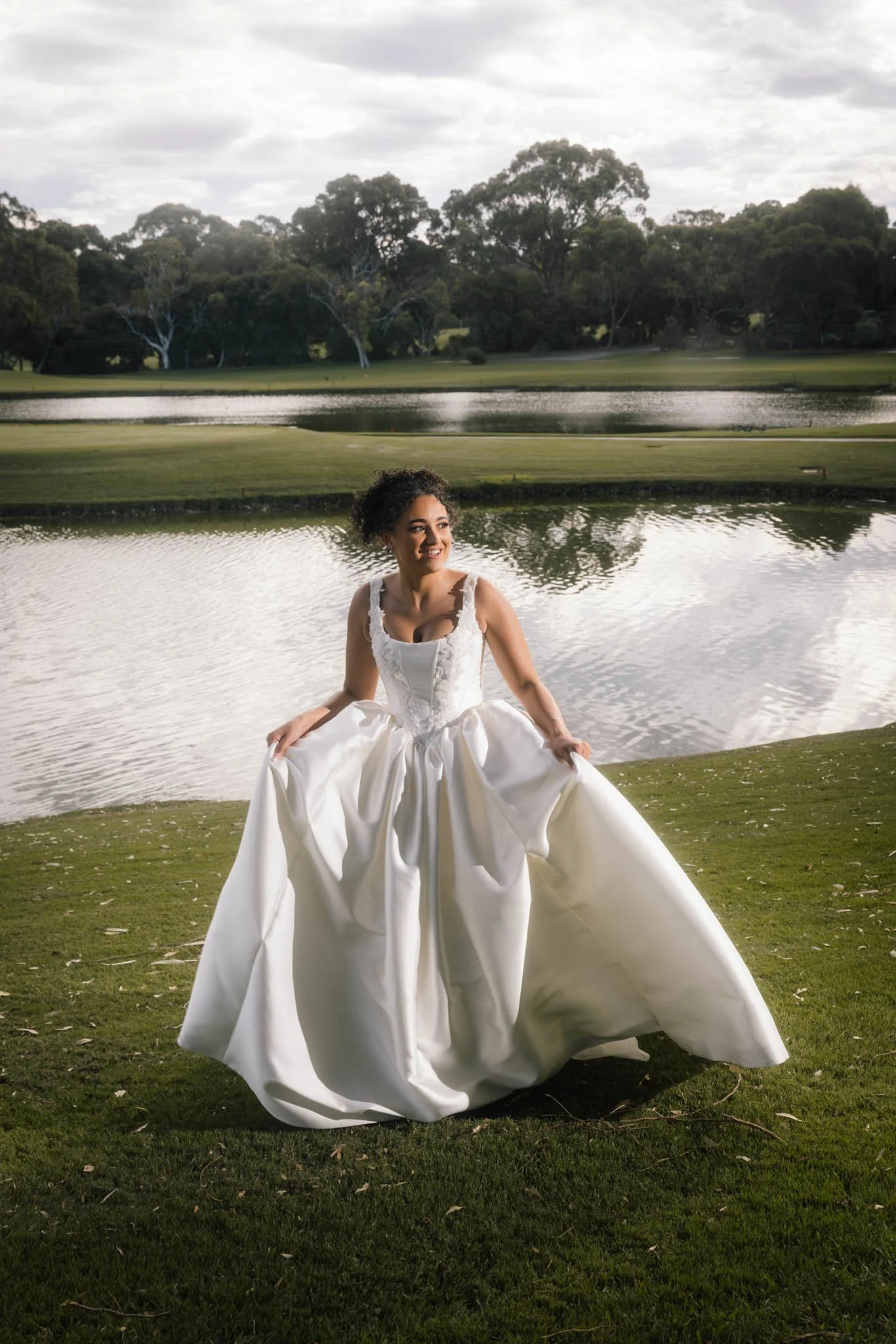 Bride in a white ball gown spreading her skirt and smiling beside the lake at Joondalup Resort Perth, with native Australian trees reflected in the water behind her, photographed by James Di Stefano