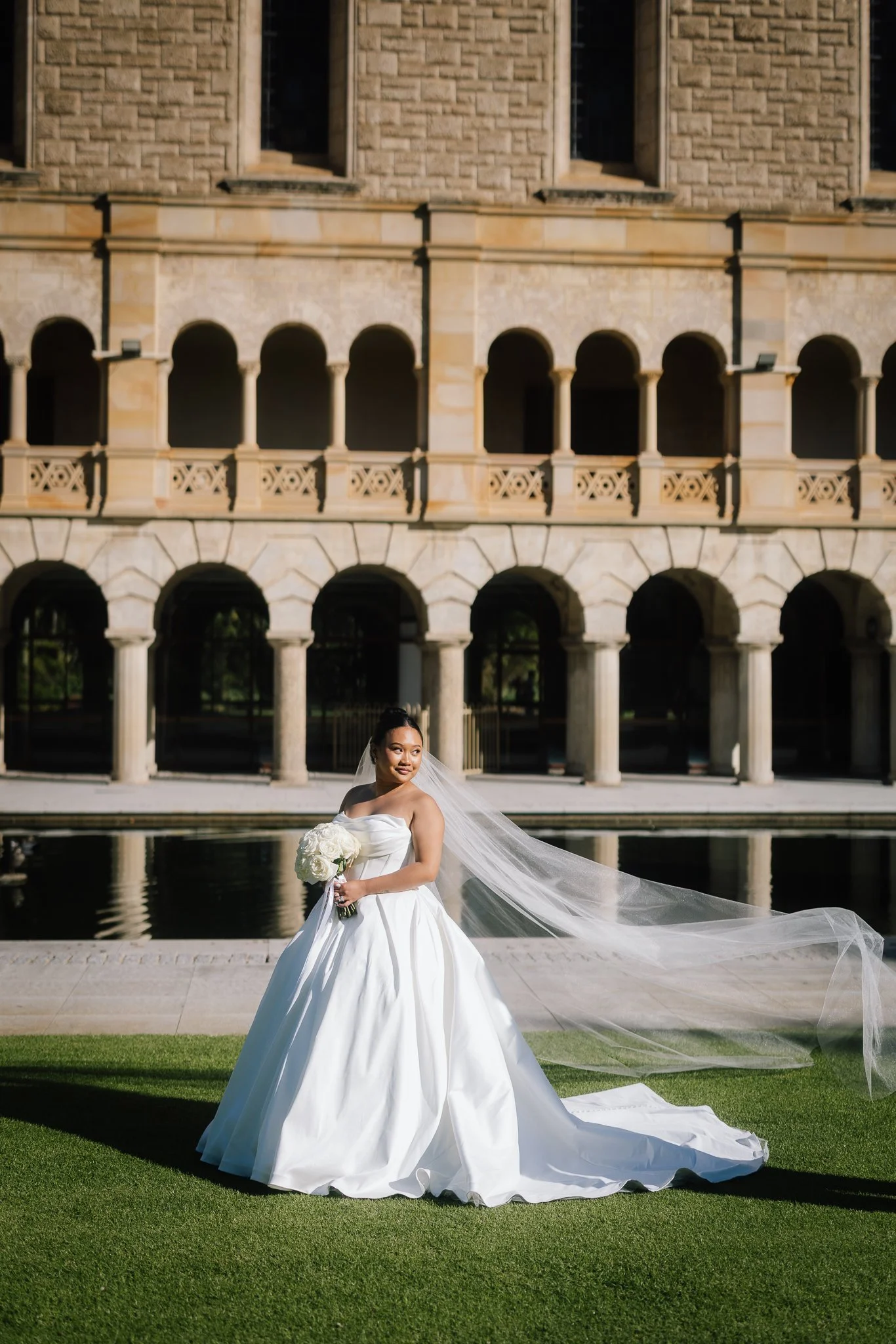 A bride's cathedral veil billows in the breeze as she stands before the UWA Sunken Garden's sandstone arches.