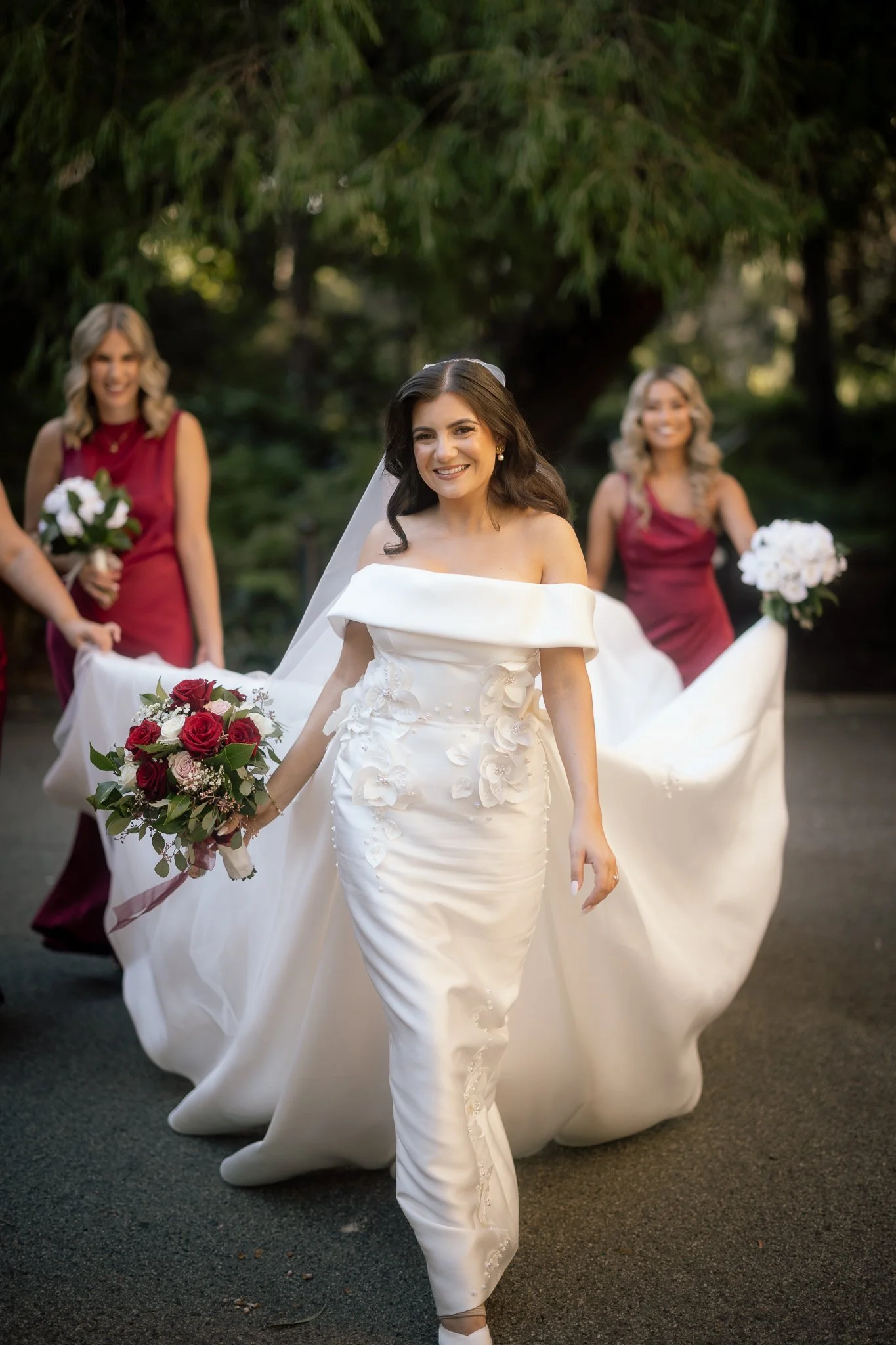 Bride in an off-shoulder satin gown with floral appliqué walks smiling as bridesmaids in burgundy carry her billowing train.
