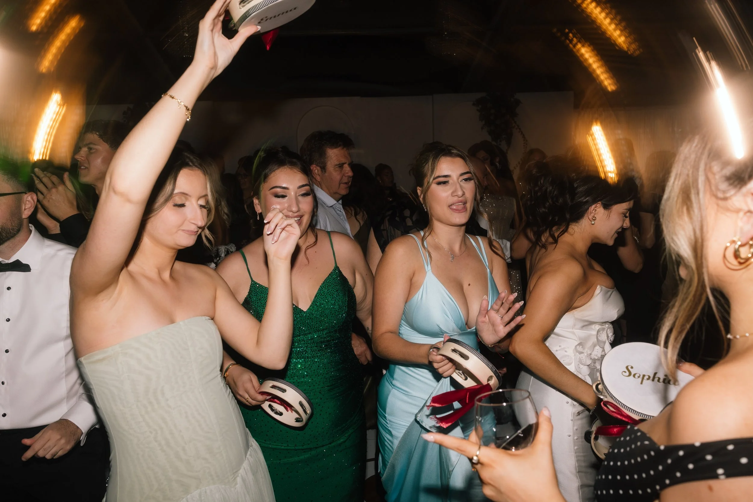 Wedding guests in green and blue gowns dance and laugh on a lively reception dance floor, holding personalised tambourines