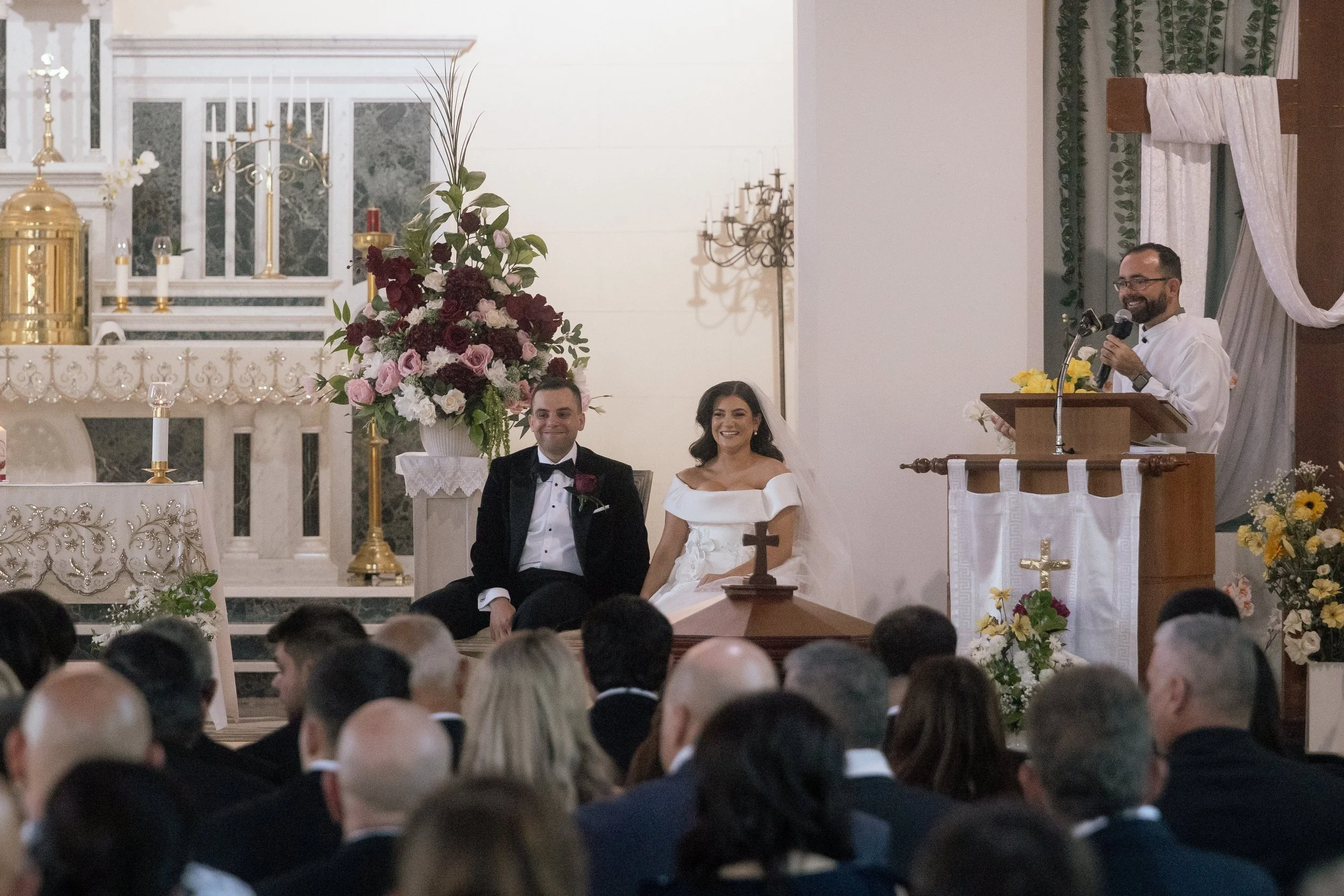 Bride and groom smile during a Catholic church ceremony in Perth, framed by lush burgundy and blush floral arrangements