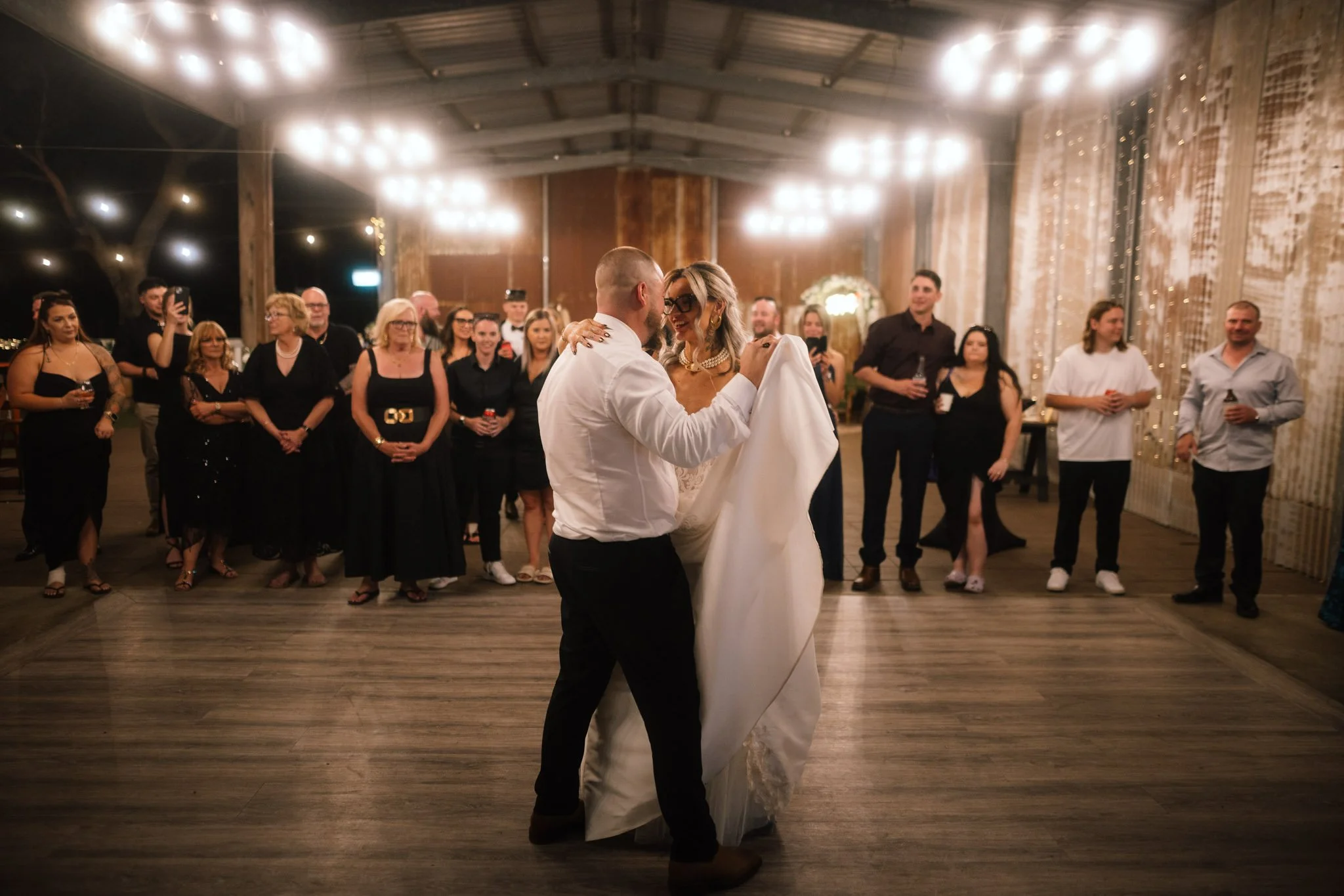 A wide-angle photograph of the bride and groom sharing their first dance inside the rustic barn wedding reception venue at Buckland Estate, surrounded by wedding guests under glowing circular chandeliers