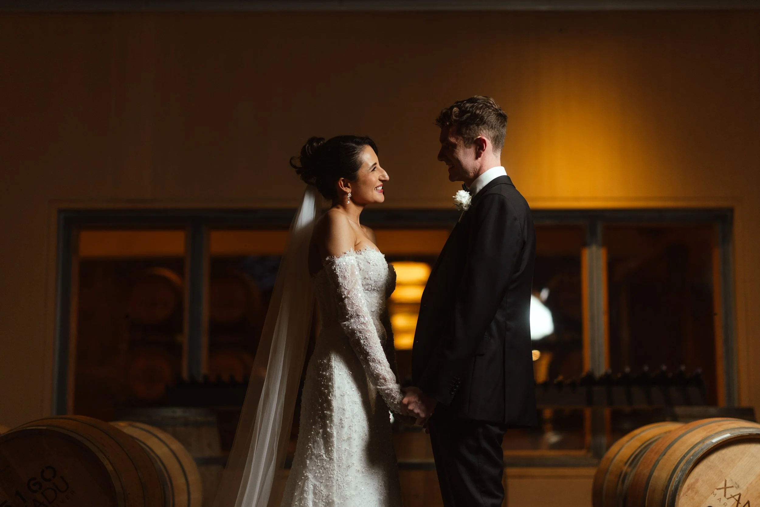 Bride in a long-sleeve lace gown and groom in black tie hold hands inside a warmly lit winery barrel room.