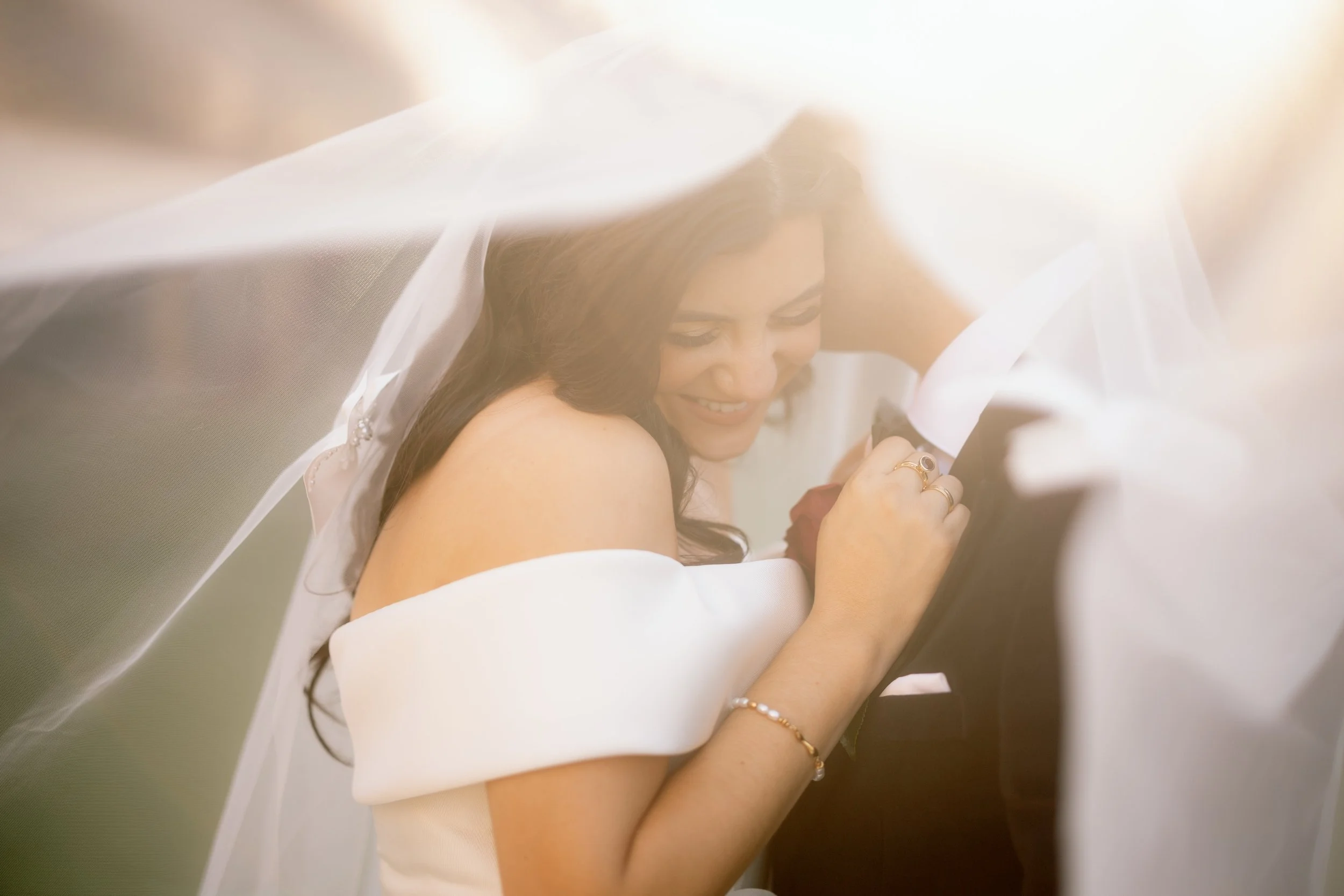 A smiling bride in an off-shoulder gown embraces her groom beneath a billowing veil in golden cinematic light.