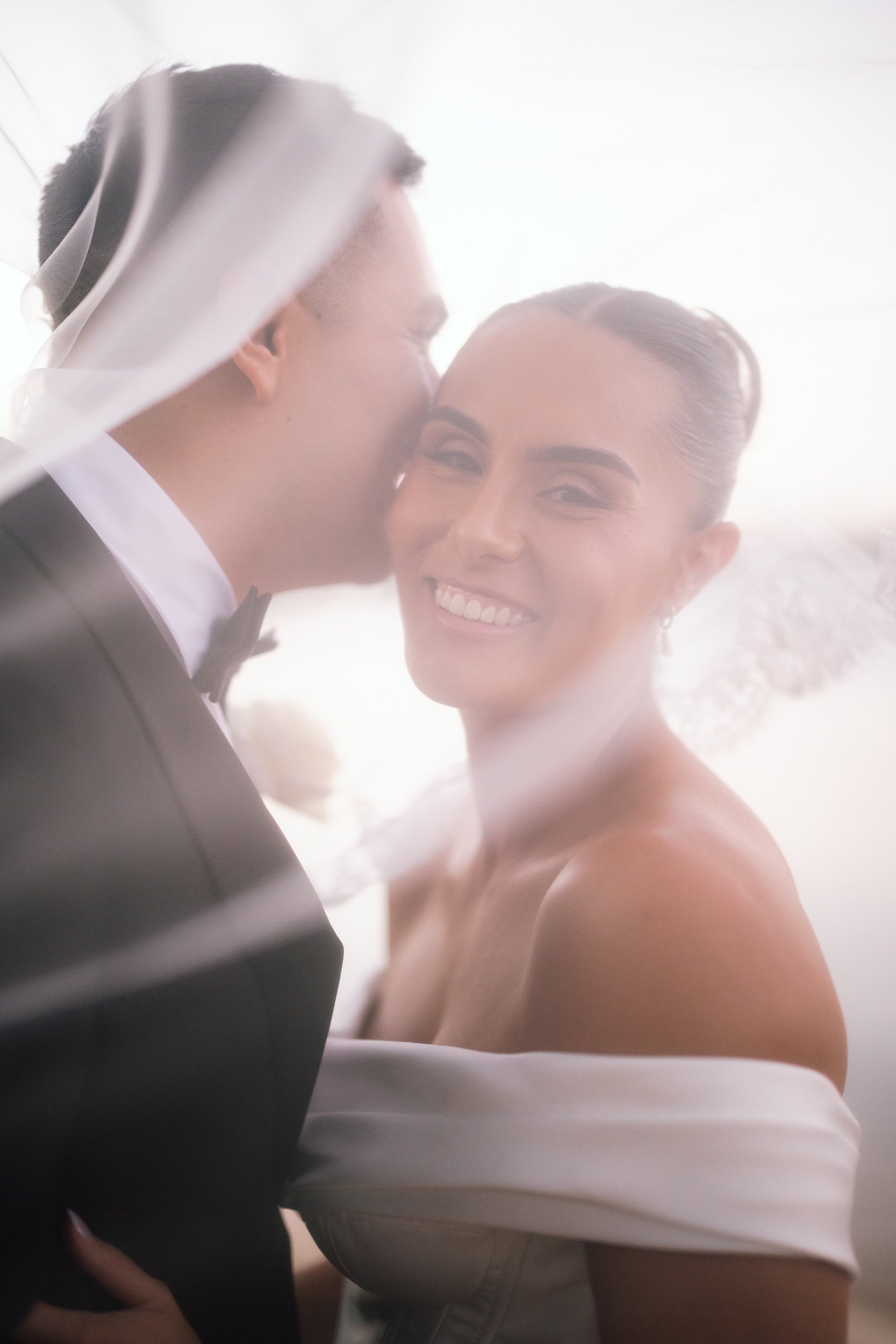 Groom kissing his bride's cheek through a billowing veil as she smiles in an off-shoulder gown during their wedding