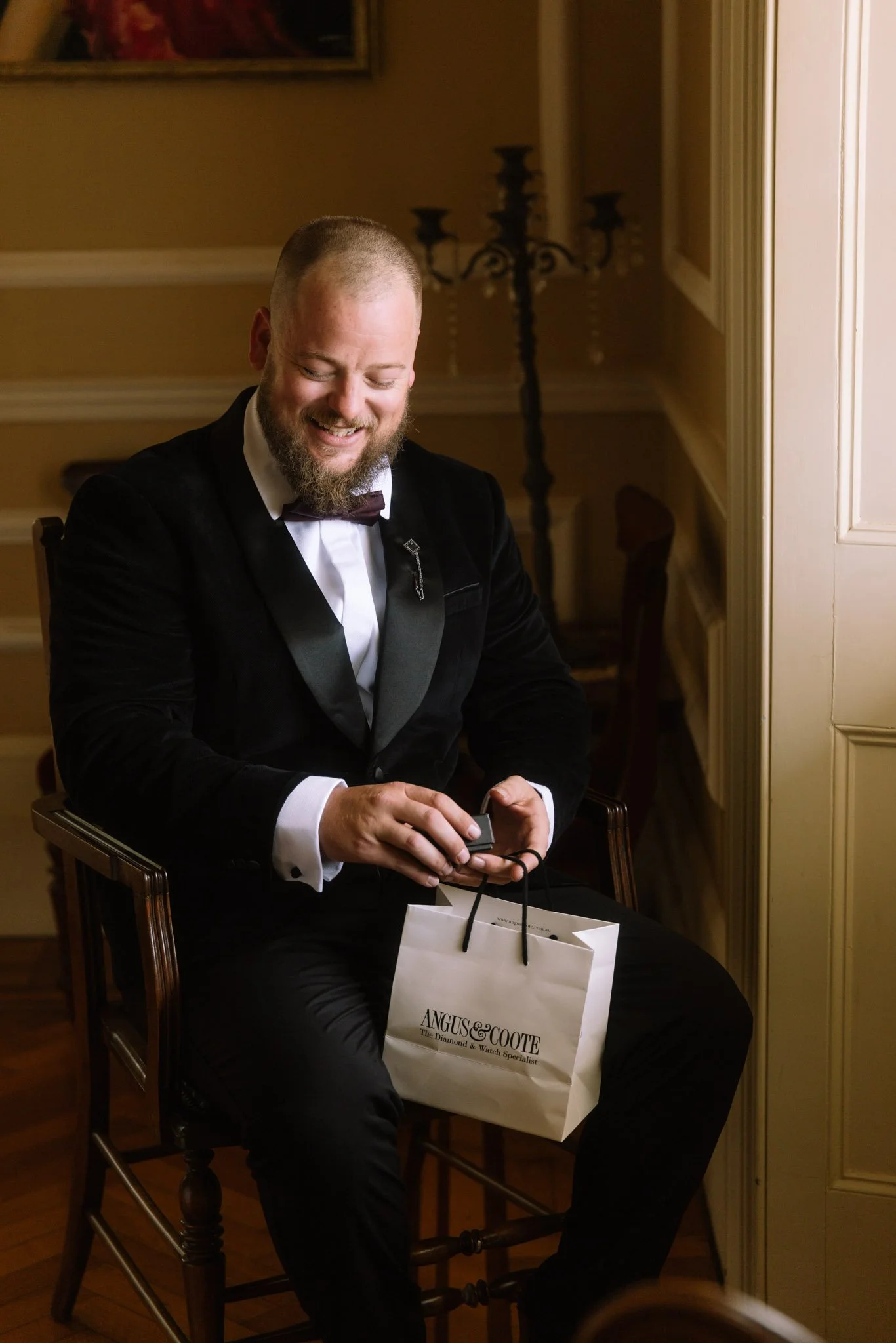 A groom smiling while opening an Angus & Coote wedding day morning gift, seated in a vintage wooden armchair inside the Buckland Estate luxury manor house.