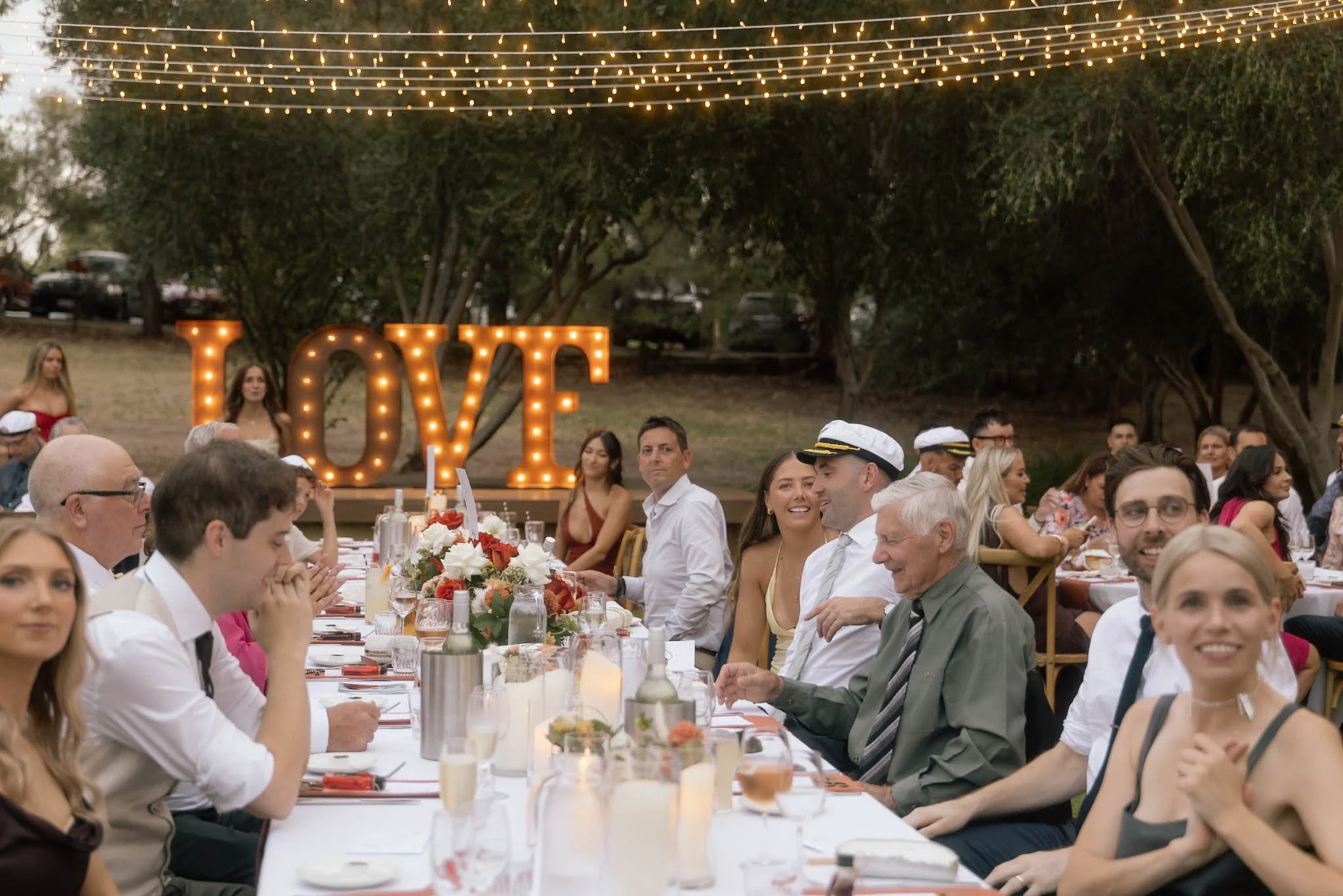 Guests seated at a long outdoor reception table at The Mallard Duck Swan Valley Perth, with glowing LOVE marquee letters and festoon lights strung through the trees overhead, photographed by James Di Stefano