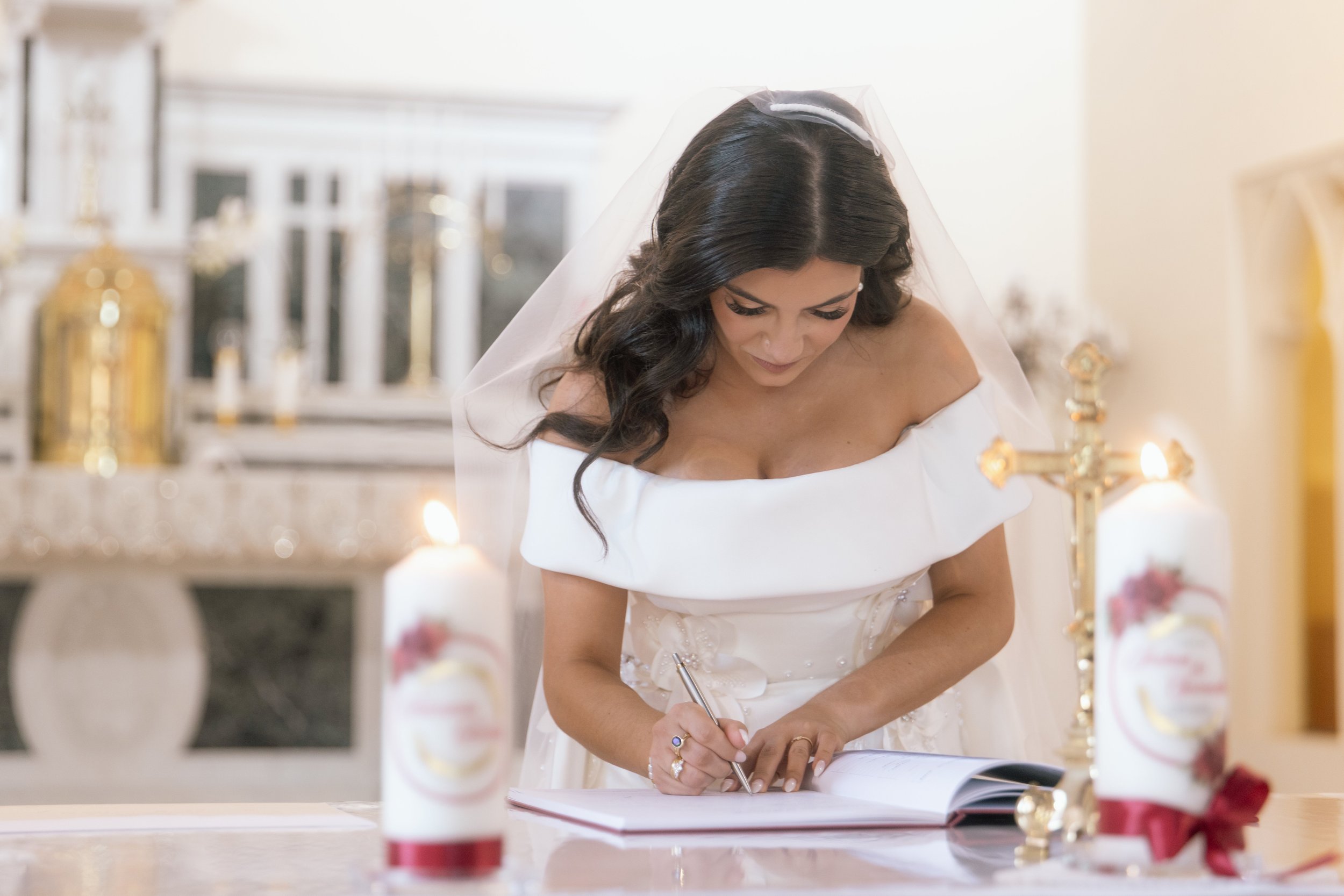 A bride in an off-shoulder gown and sheer veil signs the marriage register by candlelight during a church ceremony.