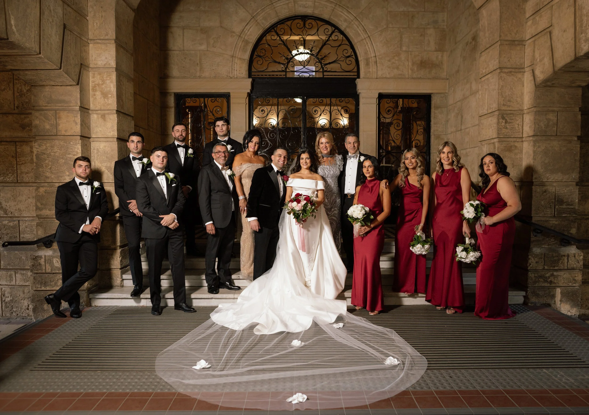 Bride in off-shoulder gown with cathedral veil, groom in black tuxedo, and wedding party in crimson and black posed on sandstone