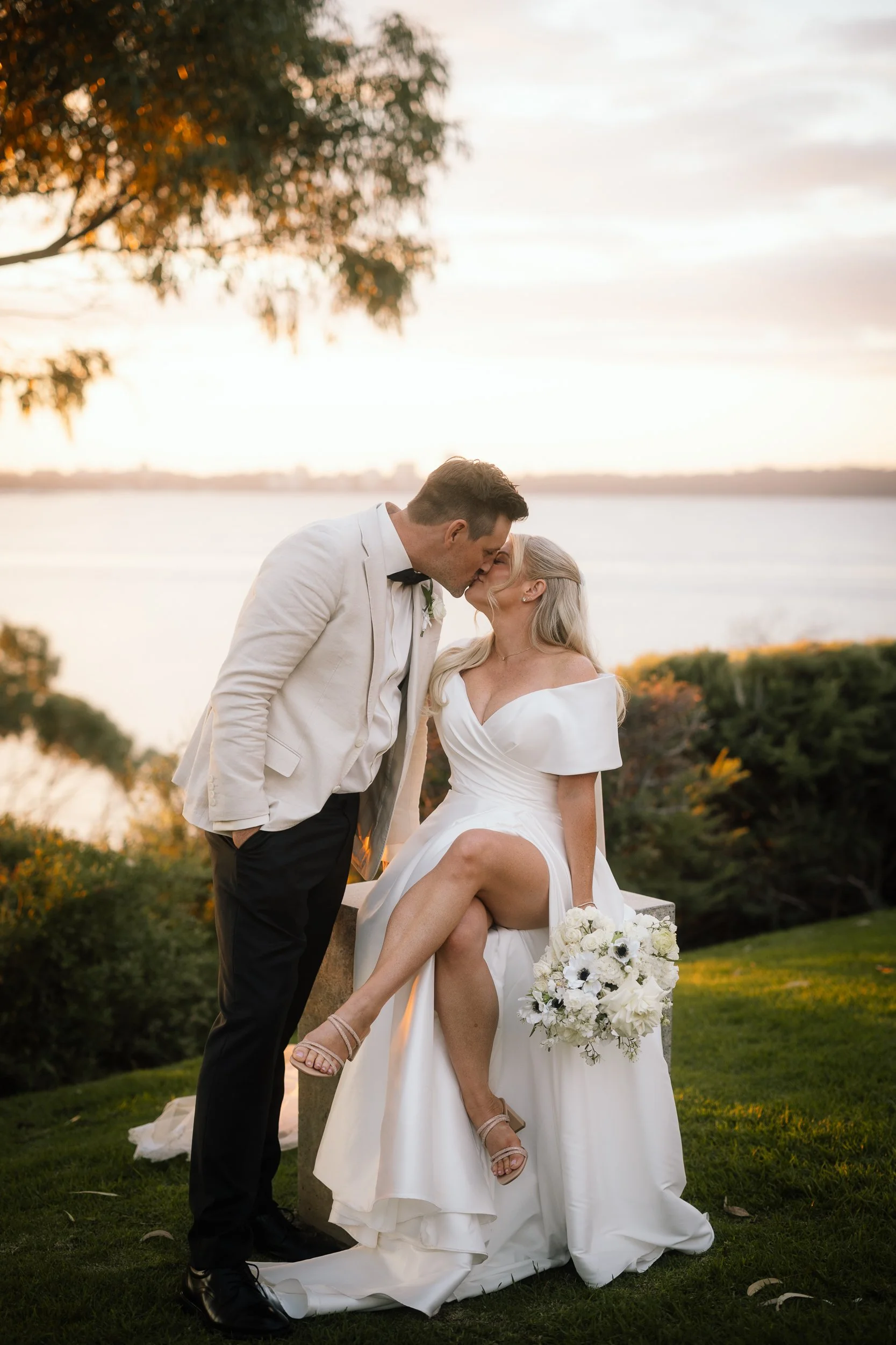 A bride in an off-shoulder gown and her groom share a golden-hour kiss beside a calm riverside waterway, her white anemone