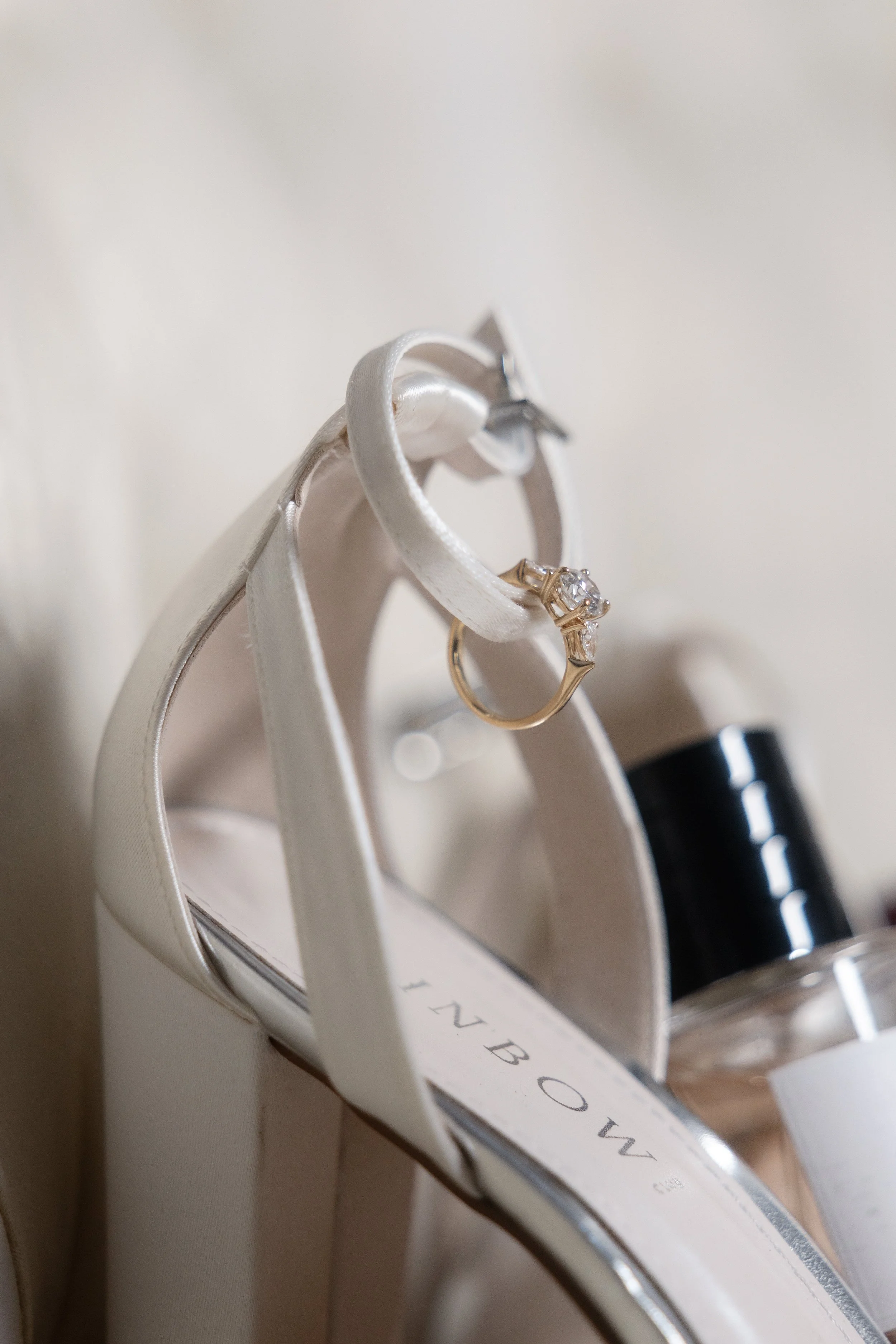A gold diamond engagement ring draped over a white satin Inbow bridal heel, styled with a perfume bottle in an editorial flatlay