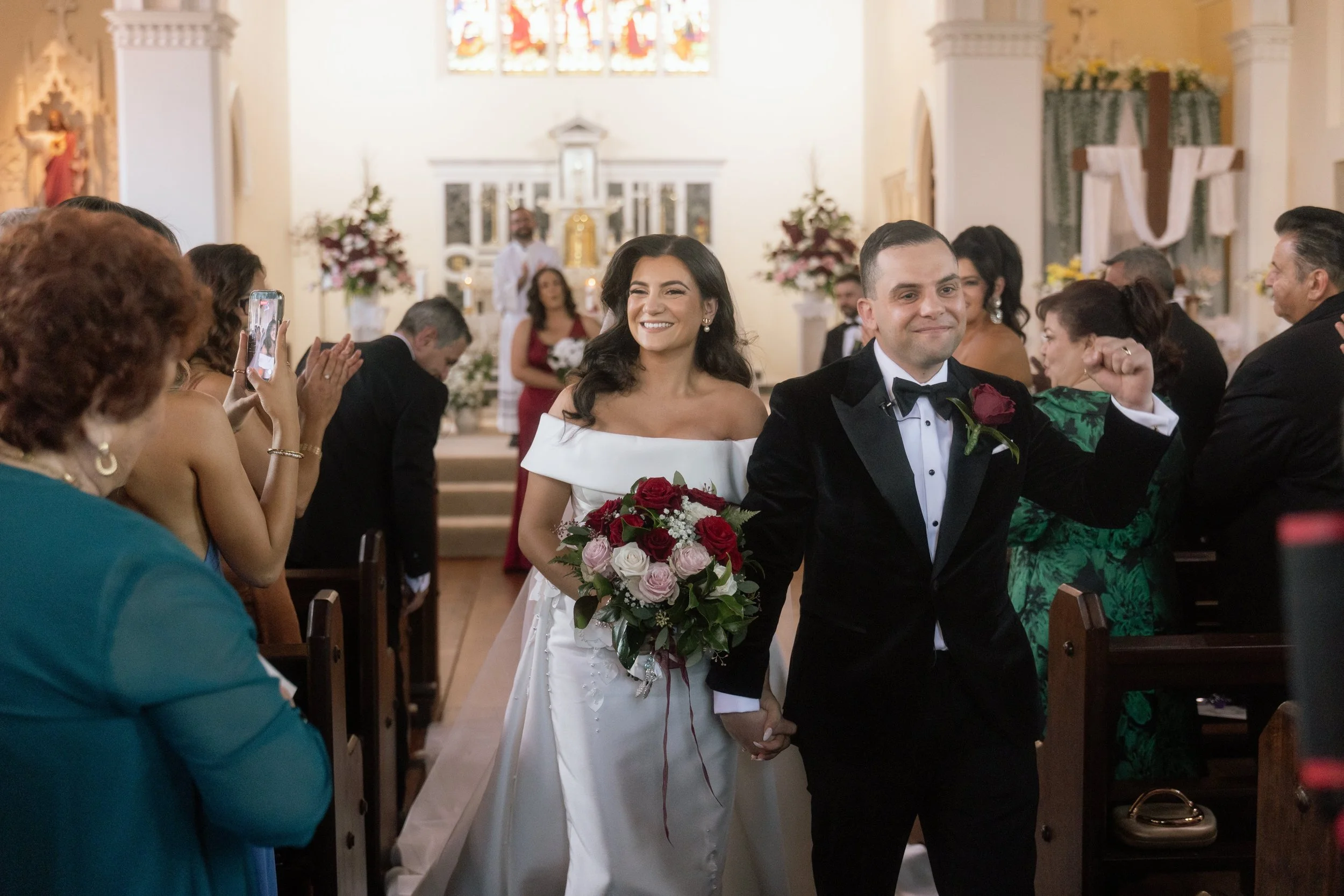 A beaming bride in an off-shoulder gown carries a red and blush bouquet as she and her groom walk back down the church aisle