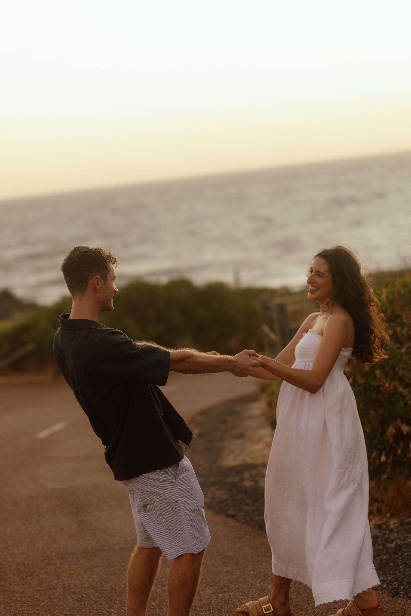 A couple laugh and spin holding hands on a coastal road at Perry's Paddock during a golden hour pre-wedding shoot.