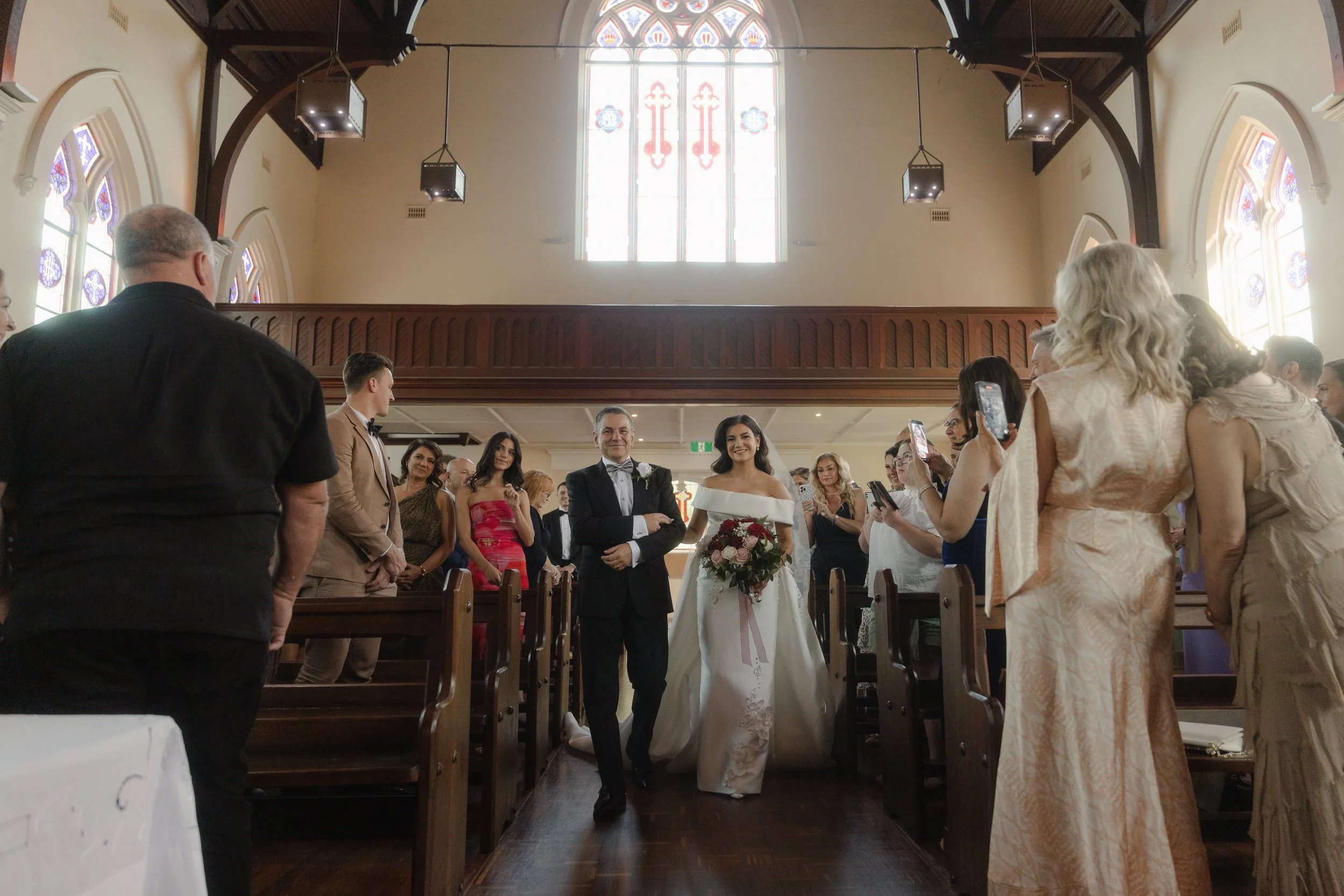 A radiant bride in an off-shoulder gown walks arm-in-arm with her father down a sunlit church aisle lined with wooden pews