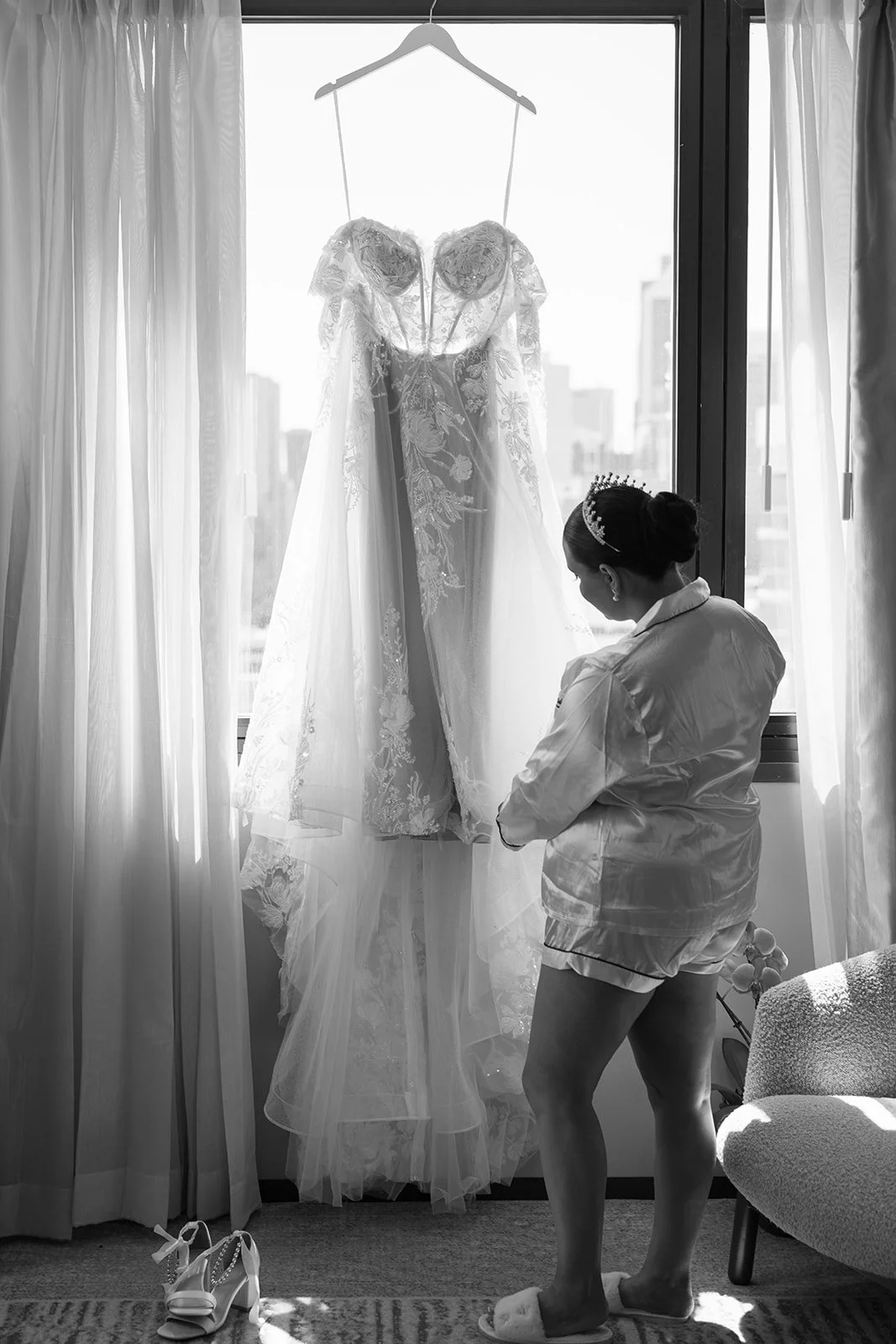 Black and white photograph of a bride admiring her detailed lace wedding gown hanging against a large city-view window at Pan Pacific Perth hotel, captured by Perth wedding photographer James Di Stefano Photo and Video.