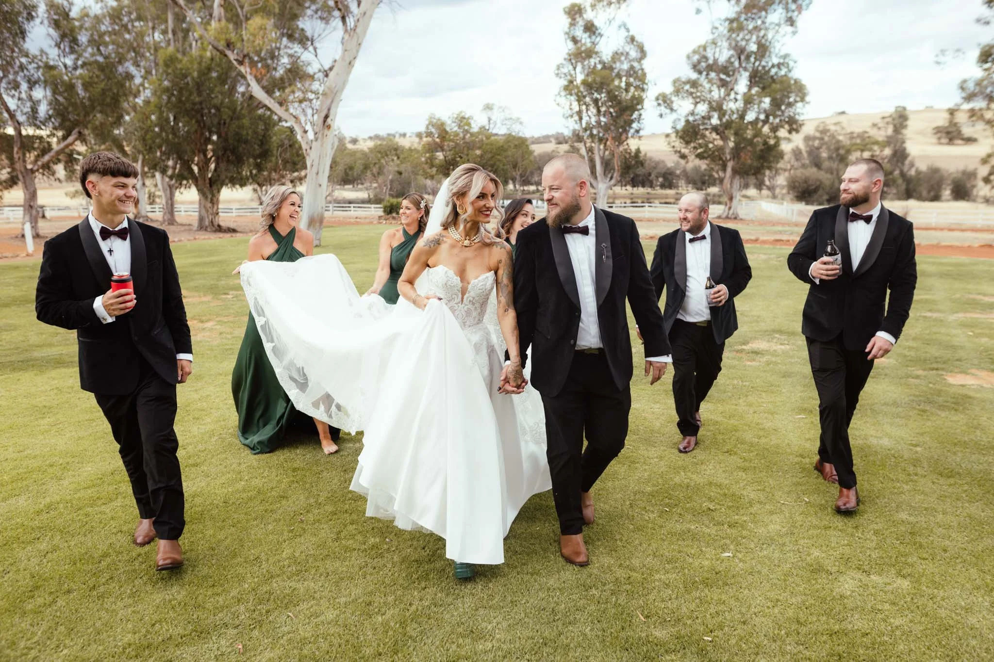 A candid and joyful photograph of a bridal party walking together across the expansive lawns at Buckland Estate. The bride and groom hold hands while bridesmaids in emerald green dresses and groomsmen in sleek black tuxedos laugh around them