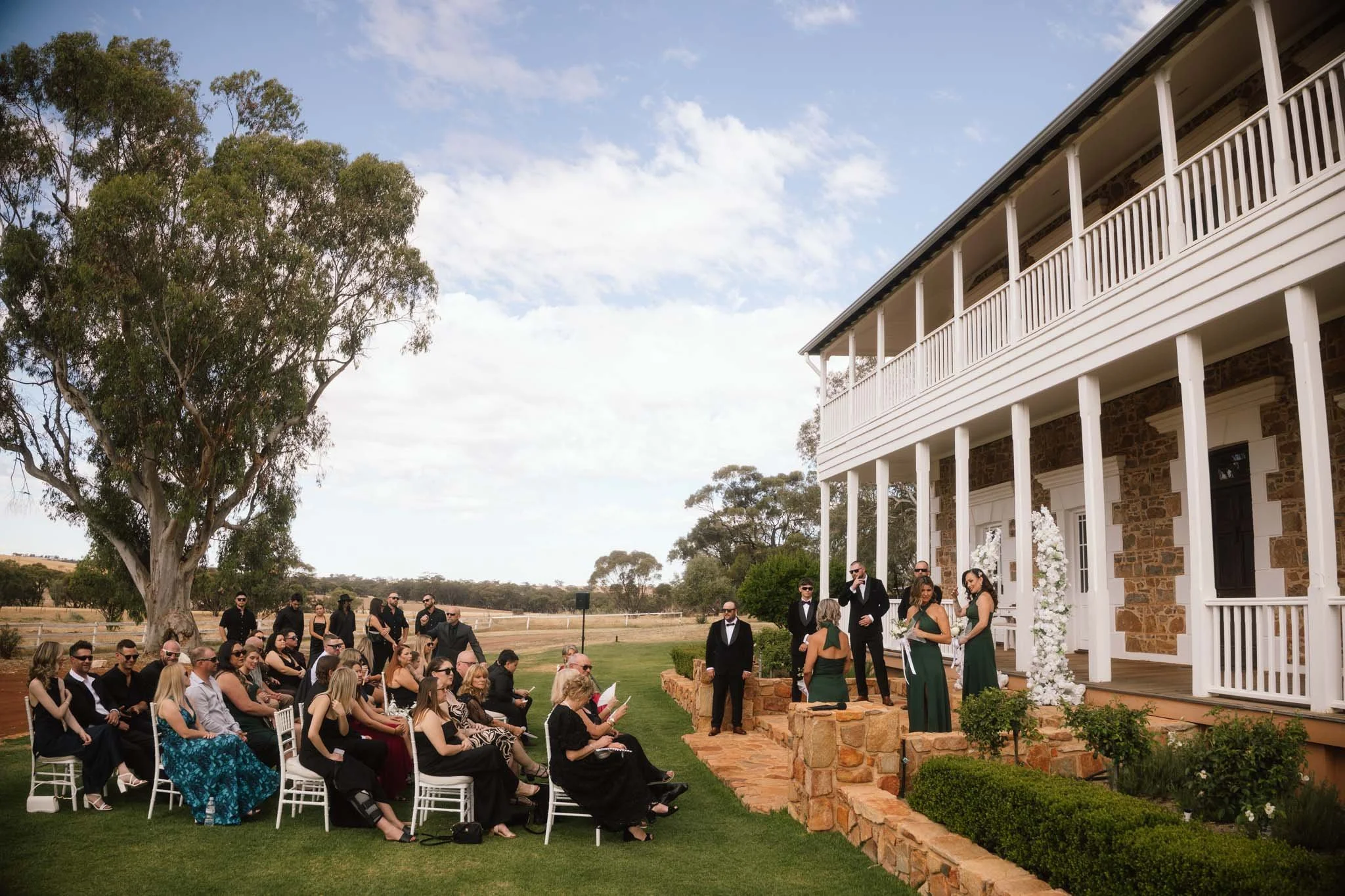 A wide photograph capturing the outdoor ceremony lawn at Buckland Estate, a hidden outback wedding venue near Perth. White chairs are neatly arranged facing a large mirror sign, all positioned in front of the grand heritage homestead.