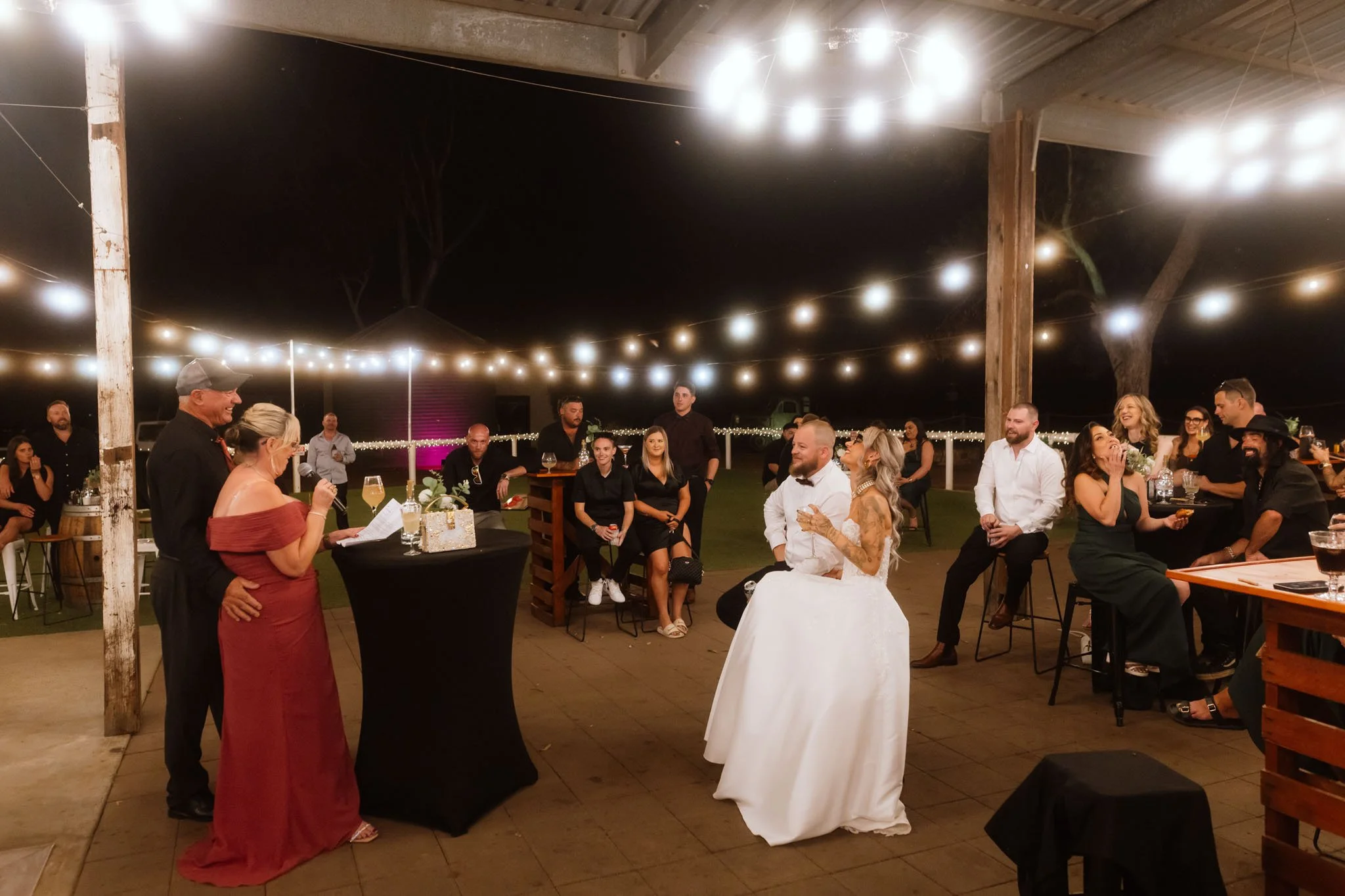 An outdoor reception scene at Buckland Estate under festoon lights, where a wedding guest gives a speech into a microphone while the bride, groom, and bridal party look on laughing