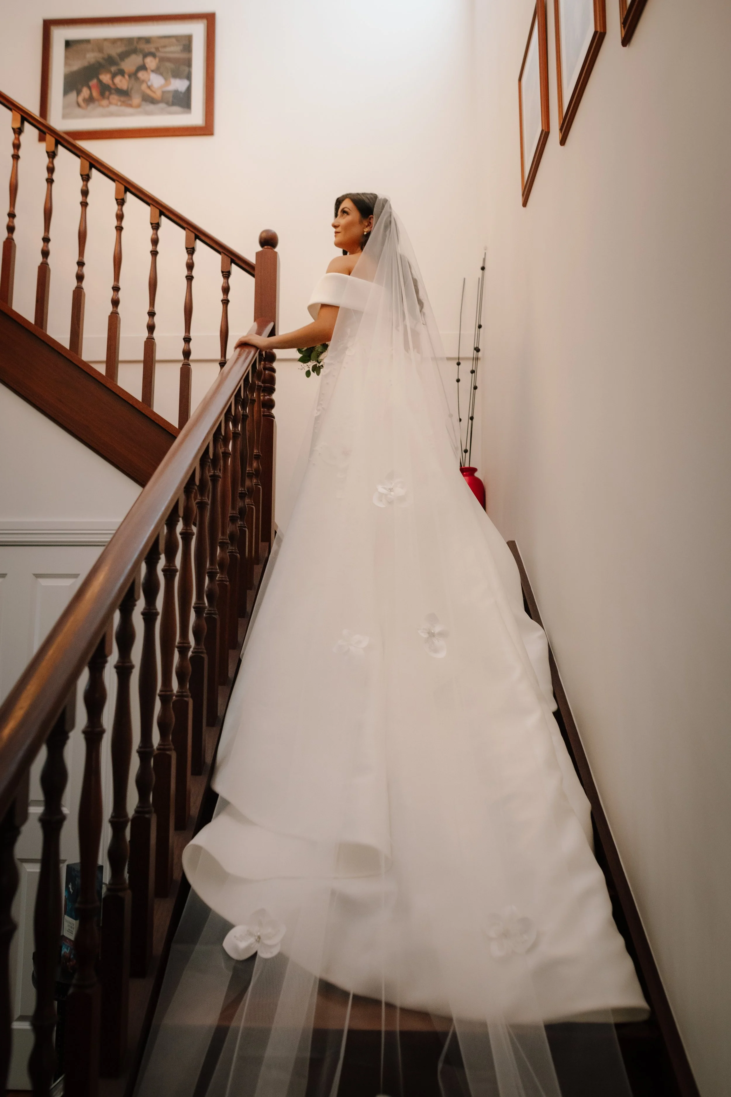 A bride in an off-shoulder ballgown with scattered floral appliqués and a cathedral veil gazes upward on a timber staircase