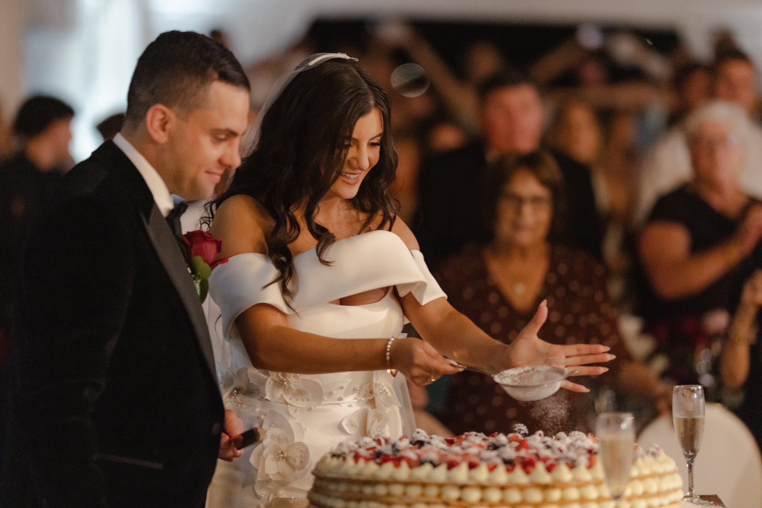Smiling bride in an off-shoulder white gown dusts icing sugar over tiered berry wedding cakes during a cinematic Perth wedding