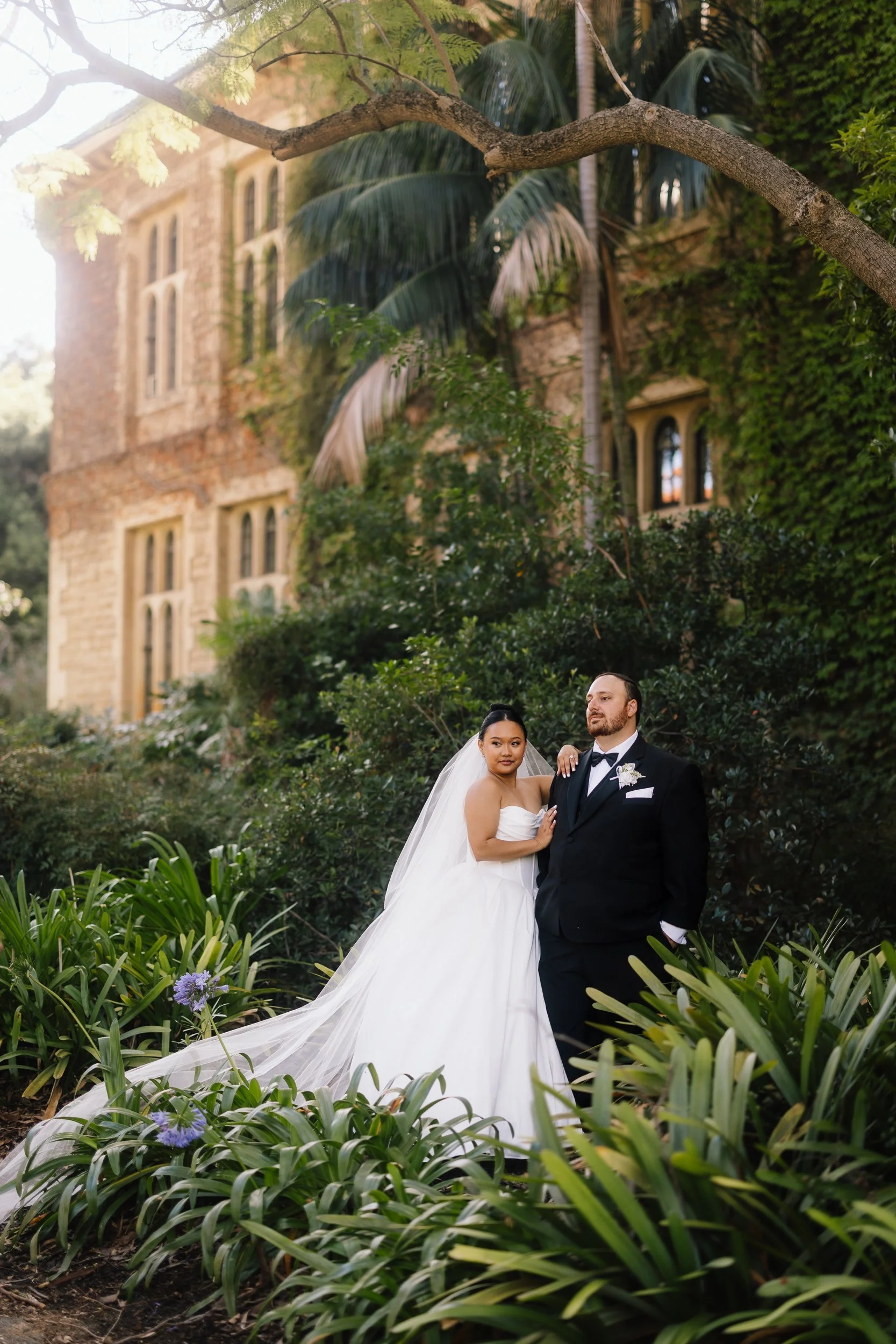 Bride in a strapless ballgown with cathedral veil stands beside her tuxedo-clad groom amid lush gardens