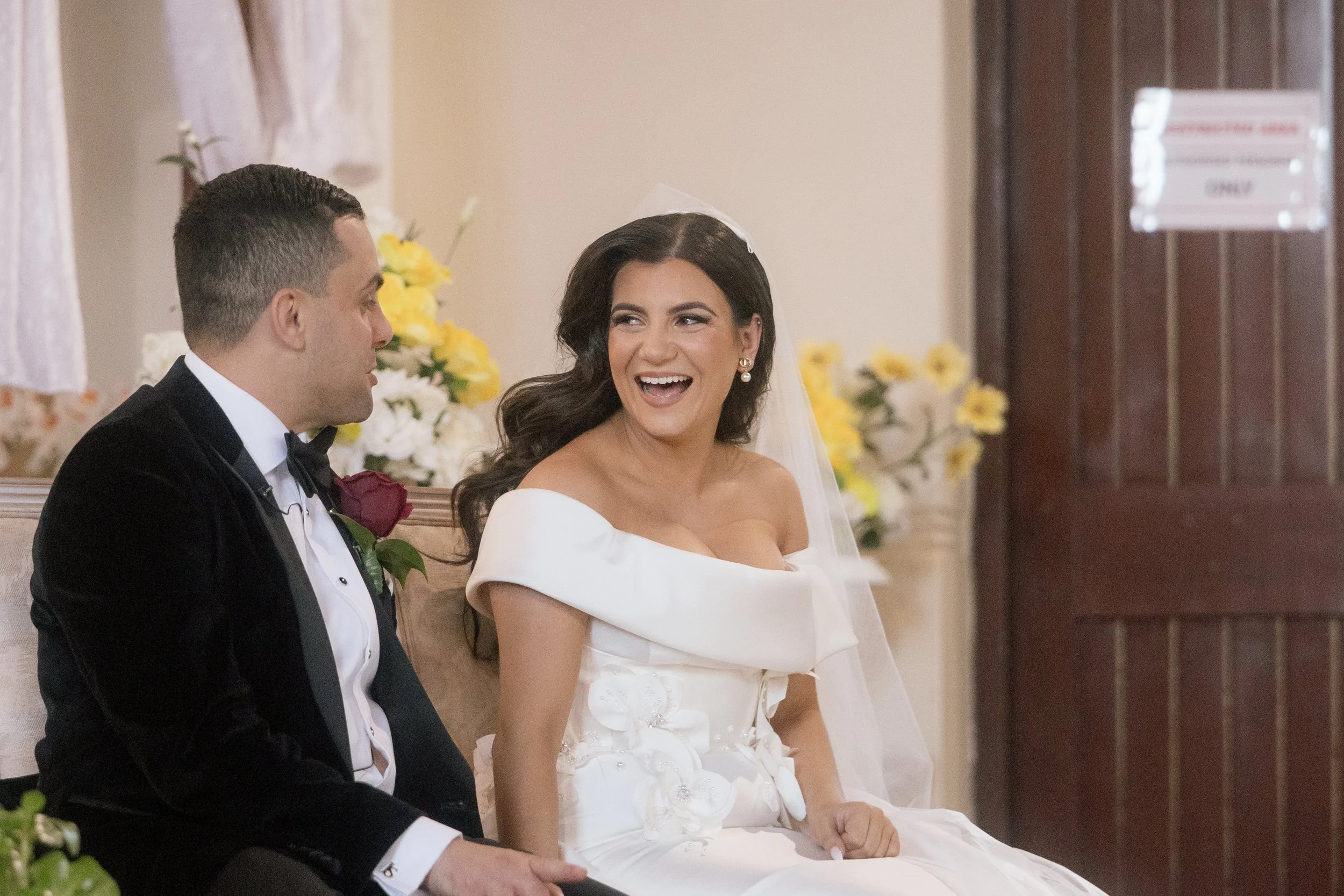 A bride in an off-shoulder gown laughs with pure joy during her wedding ceremony, her groom in a velvet tuxedo beside her.